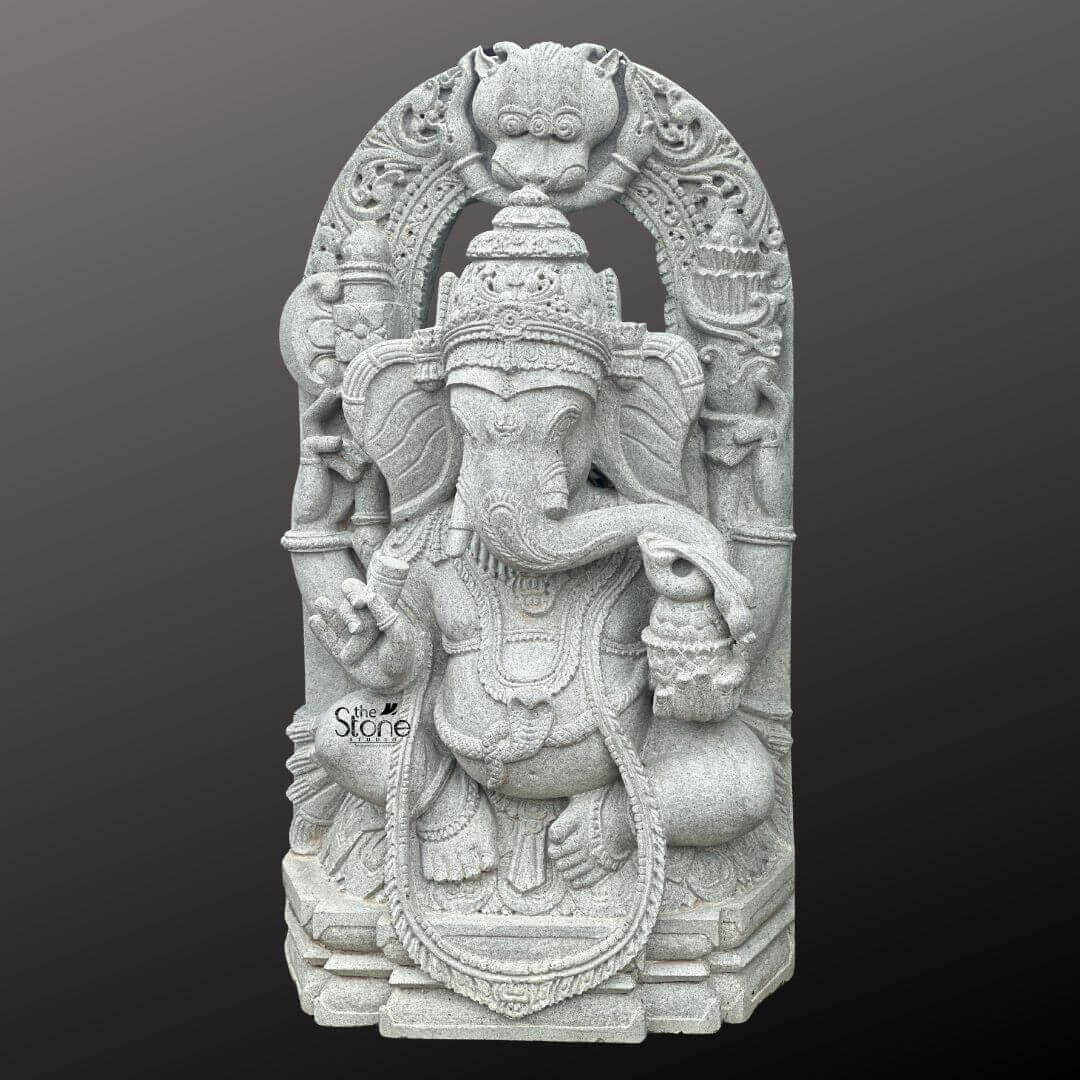 Ganesha Statue with Makara Torana 3ft 2 A magnificent 3-foot Ganesha Stone Statue with Arch intricately carved from what appears to be a light grey stone stands powerfully against a dark gradient background. The statue depicts Lord Ganesha in a regal seated posture exuding benevolence and wisdom. His iconic elephant head is finely sculpted complete with large ears expressive eyes and his distinctive broken tusk. He is adorned with an elaborate crown multiple layers of traditional jewelry including necklaces armlets and a sacred thread draped across his chest all rendered with exceptional detail in the light grey stone. Ganesha is portrayed with four arms holding his customary attributes: a modak in one hand an axe or goad in another and his lower right hand extended in a gesture of blessing offering reassurance and protection. The intricate details on his garments the patterns on his halo and the careful sculpting of his pot belly are all clearly visible showcasing the artistry of the carving. The light grey stone emphasizes these textures and details lending a serene and timeless elegance to the deity while giving the impression of an ancient weathered artifact. The statue is dramatically framed by a grand ornate arch which features exquisite mythical creature motifs and intricate traditional carvings along its entire span and supporting pillars. The top of the arch often features a Kirtimukha a fierce lion-like face a common protective motif. This entire elaborate structure rests on a multi-tiered base also carved from the same light grey stone providing stability and enhancing its grand temple-like presence. This 3-foot Ganesha Stone Statue with its impressive arch is an exquisite example of religious artistry designed to bring blessings peace and aesthetic grandeur to any space, perfectly aligning with an article focusing on such a revered Ganesha sculpture.