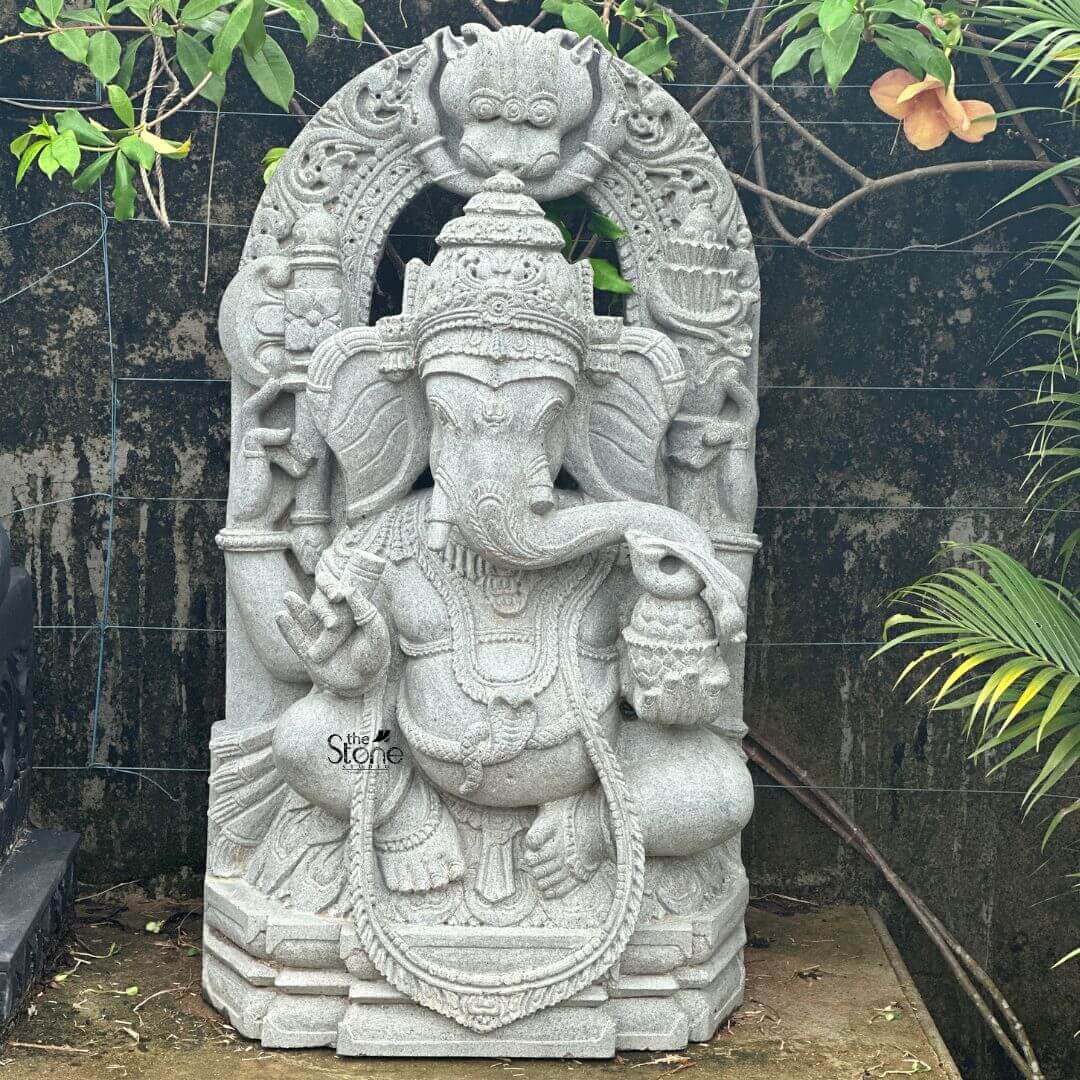Ganesha Statue with Makara Torana 3ft 3 A magnificent 3-foot Ganpati Stone Statue intricately carved from what appears to be a light grey stone is beautifully positioned in an outdoor setting ideal as a Ganpati Stone Statue for Home Temple. The statue depicts Lord Ganesha in a regal seated posture exuding benevolence wisdom and an aura of divine grace. His iconic elephant head is finely sculpted complete with large ears expressive eyes and his distinctive broken tusk delicately poised to write. He is adorned with an elaborate crown multiple layers of traditional jewelry including necklaces armlets and a sacred thread draped across his chest all rendered with exceptional detail in the light grey stone. Ganesha is portrayed with four arms holding his customary attributes a modak sweet dumpling in one hand an axe or goad in another and his lower right hand extended in a gesture of blessing offering reassurance and protection. The intricate details on his garments the patterns on his halo and the careful sculpting of his prominent pot belly are all clearly visible showcasing the high artistry of the carving. The light grey stone emphasizes these textures and details lending a serene and timeless elegance to the deity and suggesting a weathered ancient feel that would be magnificent in a home temple. The statue is dramatically framed by a grand ornate arch that features exquisite mythical creature motifs and intricate traditional carvings along its entire span and supporting pillars. The top of the arch features a Kirtimukha a fierce lion-like face a common protective motif. This entire elaborate structure rests on a multi-tiered base also carved from the same light grey stone providing stability and enhancing its grand temple-like presence. This 3-foot Ganpati Stone Statue with its impressive arch is an exquisite example of religious artistry designed to bring blessings peace and aesthetic grandeur to any home temple or sacred space perfectly aligning with an article focusing on such a revered Ganpati sculpture.