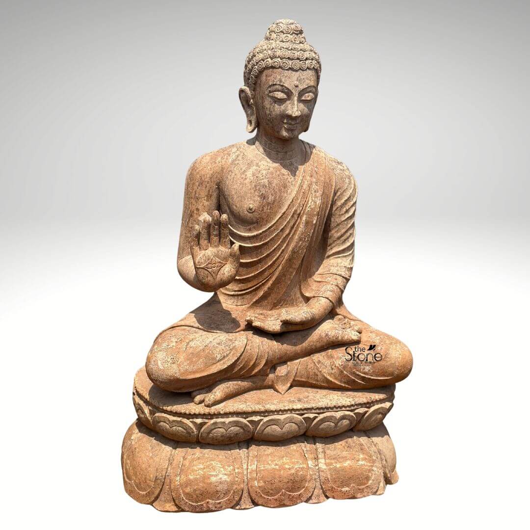 Sandstone Buddha Statue in Abhaya Mudra 3ft 5 A magnificent 3-foot Sandstone Buddha statue serving as an ideal Garden Buddha Statue stands prominently against a clean light grey background radiating peace and reassurance. The statue carved from a warm reddish-brown sandstone depicts Buddha in a serene meditative posture seated in the full lotus position symbolizing deep concentration and spiritual enlightenment. His right hand is raised to shoulder height with the palm facing outwards fingers extended upwards forming the Abhaya Mudra the gesture of fearlessness blessing and protection. His left hand rests gently in his lap palm upwards signifying contemplation and receptivity. The natural earthy hue and weathered texture of the sandstone give the sculpture an authentic ancient feel perfectly suited for an outdoor garden environment. The Buddha's facial features are exquisitely carved radiating profound peace and wisdom. His eyes are gently closed or cast downwards in contemplation and a subtle serene smile graces his lips. His head is adorned with tightly coiled hair culminating in the ushnisha the cranial bump symbolizing his supreme spiritual knowledge and enlightenment. The flowing drapery of his robes is sculpted with soft elegant folds emphasizing the texture of the sandstone and contributing to the statue's graceful and tranquil form. The rustic worn appearance of the sandstone enhances its charm and allows it to blend seamlessly into a natural garden setting gaining character over time. The Buddha is seated upon a beautifully sculpted double-lotus pedestal with each petal intricately carved symbolizing purity spiritual growth and the emergence of enlightenment from the mundane. The robust and detailed base supports the statue with dignit making it a focal point. This 3-foot Sandstone Buddha statue with its clear depiction of the Abhaya Mudra and durable sandstone material is an exquisite example of religious artistry designed to bring blessings, peace and aesthetic beauty to any garden patio or outdoor space aligning perfectly with an article focusing on Garden Buddha Statues in Sandstone.