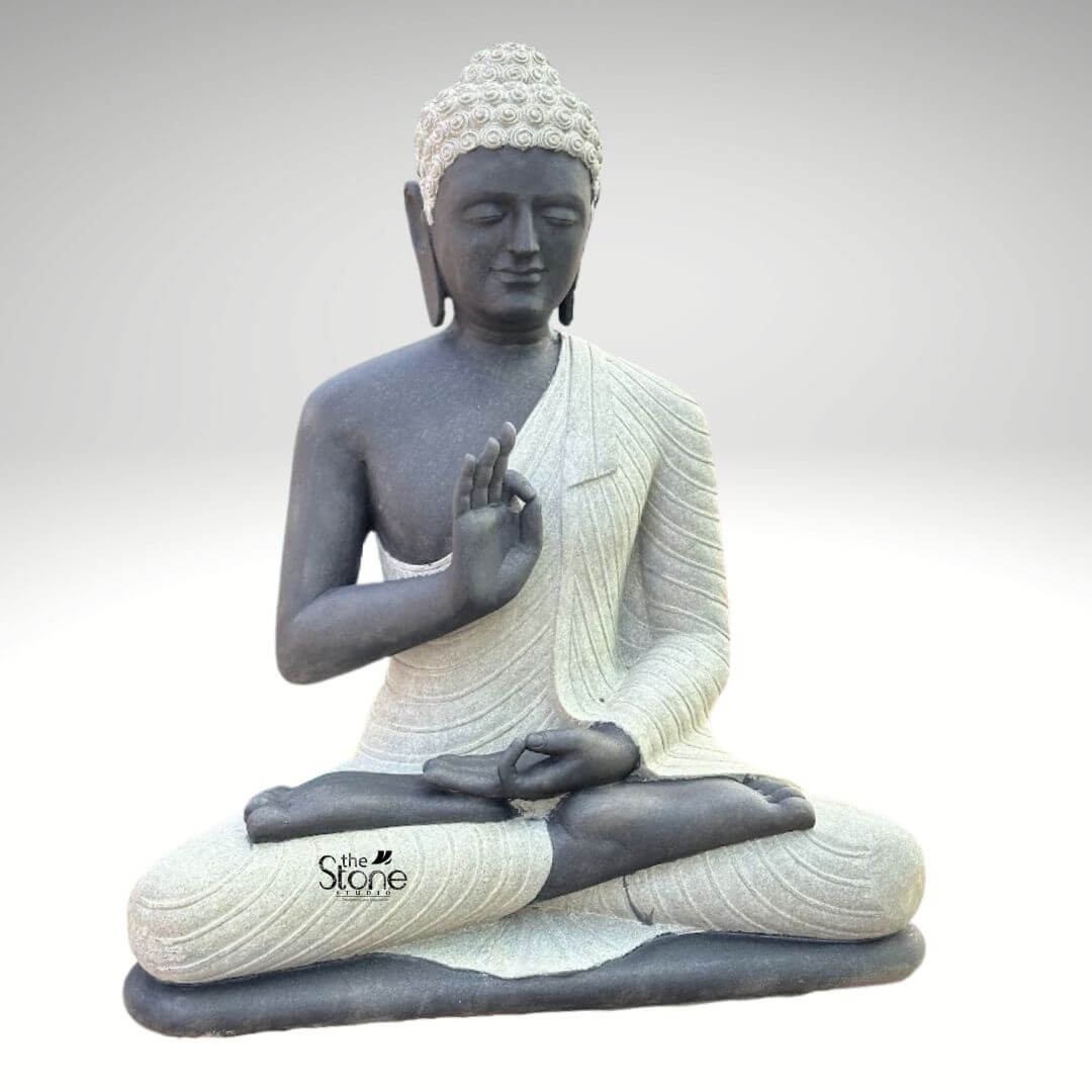 Two Tone Buddha Stone Statue for Home 3ft 2 A serene hand-carved Blackstone Buddha statue sits in a traditional meditative pose presented in a clean studio setting against a soft grey background. The three-foot-tall statue showcases a striking two-tone finish with the Buddha's body and head carved from a deep dark blackstone, exhibiting a smooth polished texture that highlights the rich dark tones. This contrasts beautifully with his monastic robes which are rendered in a contrasting lighter grey or off-white stone featuring a subtle granular texture. This distinct separation emphasizes the form of the robes as they drape over his crossed legs and shoulders adding depth and visual interest to the sacred art. The Buddha is depicted seated with his legs crossed in a full lotus position on a simple integrated dark blackstone base. His hands are gracefully positioned in the vitarka mudra symbolizing discussion and the transmission of teachings with the right hand raised and the thumb and index finger forming a circle while the left hand rests in his lap. His facial expression is calm and contemplative with downcast eyes a gentle smile and tightly curled hair leading to a cranial ushnisha all rendered with precise craftsmanship that honors traditional iconography. The overall composition evokes a sense of peace tranquility and spiritual harmony. This three-foot Blackstone Buddha statue is an ideal illustration for an article discussing a Garden Buddha Statue demonstrating how such a piece can transform an outdoor space into a peaceful sanctuary. Its substantial size ensures it functions as a commanding focal point while the natural stone texture harmonizes beautifully with surrounding plant life. The traditional design brings a sense of sacred history and calm to any garden landscape while the durable blackstone construction ensures it is both a beautiful and a meaningful addition to the home garden. The detailed carving and harmonious integration of elements highlight its value as a piece of sacred art in a modern landscape.