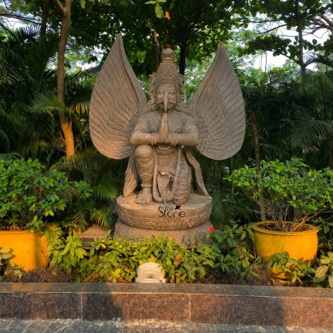 Grey Stone Garuda Statue 5.5ft 3 A full-length three-quarter view photograph presents a magnificent 5.5-foot-tall Garuda statue meticulously hand-carved from a single block of grey granite. The statue is the central focal point of an outdoor garden setting elevated upon a robust circular lotus-carved pedestal base. Garuda, the mythical bird-deity is depicted in a reverent kneeling posture with both hands pressed together in the Anjali Mudra a gesture of prayer and devotion held near his chest. His face is carved with intricate detail featuring an eagle-like beak defined eyes, and a traditional crown with a coiled serpent motif. His powerful form is adorned with deeply carved traditional ornaments including layered necklaces armbands, bracelets and a sacred thread. A defining feature of this grey granite Garuda sculpture is its massiv fully spread wings, which rise dramatically behind him, mirroring his height and creating a sense of divine presence. Each feather is individually and delicately carved creating a texture that is both visually detailed and structurally impressive. The granite stone itself possesses a natural slightly matte finish with subtle crystalline variations in color ranging from light to medium grey giving the sculpture an authentic ancient feel. The entire form balances grace with power capturing the essence of the revered celestial mount of Lord Vishnu. This five-and-a-half-foot grey stone Garuda statue is an exemplary piece of Indian stone carving craftsmanship, making it a perfect subject for an article discussing granite sculptures. Its commanding size and detailed craftsmanship make it suitable for placement in a home temple a garden sanctuary, or as an architectural element in a larger space. The choice of durable grey granite ensures its longevity making it a timeless investment for spiritual and aesthetic appreciation. The image captures the unique iconographic elements and the high level of artistry that define such a premium stone statue.