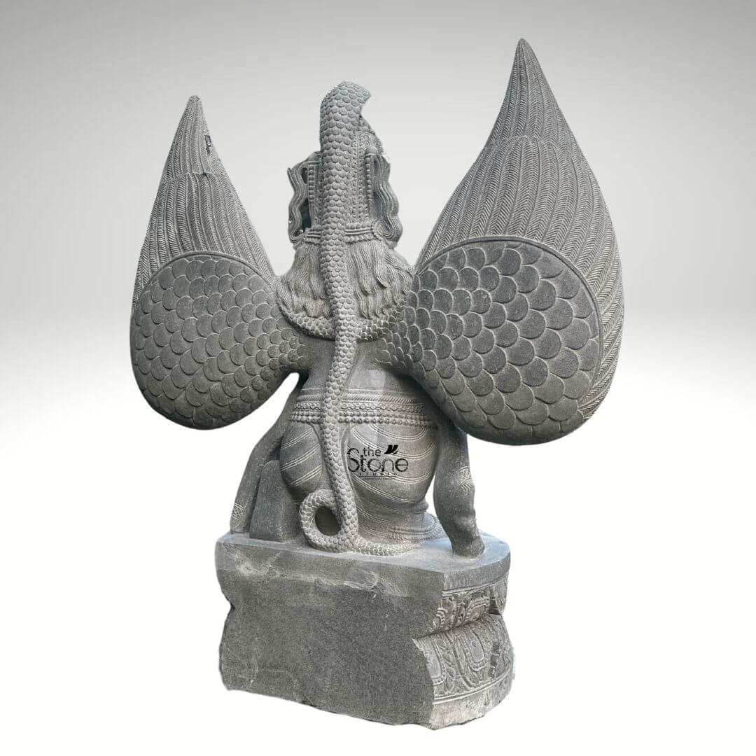 Grey Stone Garuda Statue 5.5ft 4 A detailed rear-facing photograph captures a magnificent 5.5-foot-tall Granite Garuda Idol meticulously hand-carved from a single block of natural grey granite. The sculpture presents a powerful and complex composition from behind showcasing the intricate artistry of traditional stone carving. The central figure is the mythical bird-deity Garuda, depicted with large folded wings that rise prominently their surface textured with overlapping scale-like feathers that catch the light. A long, textured serpent is coiled dramatically along the entire length of Garuda's spine and tail its scales and segmented body rendered with fine precision creating a striking contrast with the larger feather patterns. Garuda's form including his broad shoulders and defined back muscles is revealed beneath the coiled Naga and his lower body is draped in simple carved garments. The Garuda idol is elevated standing upon a robust carved rectangular stone pedestal base. The pedestal itself is adorned with decorative reliefs on its visible sides, adding to the overall grandeur of the sculpture. The grey granite material has a natural slightly matte finish with subtle, organic color variations and crystalline flecks emphasizing its authenticity and durability as an idol designed for worship or prominent display. The carving exhibits a high level of detail from the individual feather points to the segmented body of the Naga, reflecting the skill and patience of the master artisan who created it. The statue's pose is one of contained power capturing the essence of the revered celestial mount of Lord Vishnu. This five-and-a-half-foot Granite Garuda Idol is an exceptional example of traditional Indian stonework making it a perfect subject for an article discussing granite sculptures. Its large size and detailed craftsmanship ensure it will serve as a commanding and spiritual focal point in a home temple a garden sanctuary, or as an architectural element in a larger space. The symbolic significance of Garuda as a protector adds a layer of cultural and religious depth. The rear view offers a unique and detailed perspective of the statue's complex iconography particularly the dramatic depiction of Garuda with the Naga which is a key element of his legend. The durable grey granite guarantees its longevity and aesthetic appeal for years to come.