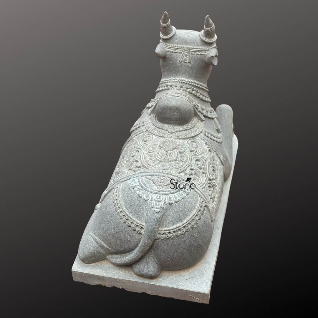 Nandikeshwara Stone Statue 45" 2 A high-angle rear three-quarter photograph captures a beautifully carved Nandi bull statue hand-hewn from a single block of dark grey blackstone. The statue depicts Nandi the sacred mount and devoted follower of Lord Shiva in a traditional serene reclining posture resting upon a simple elevated rectangular blackstone plinth. The stone has a fine-grained matte finish that emphasizes the rich deep color and intricate sculptural details. The view from the back reveals the powerful yet graceful form of the bull with careful attention paid to the musculature of the hindquarters and back. Nandi is richly adorned with traditional deeply carved ornaments that showcase the artisan's skill. A large ornate blanket covers his back featuring elaborate symmetrical floral paisley and geometric patterns in high relief. This blanket extends down the sides with intricately carved fringed borders. Around his neck and hump he wears multiple layered necklaces of varying designs including beaded chains bells and floral pendants all rendered with fine detail. The carving extends to his legs which are decorated with anklets. His horns are carefully shaped and point upwards and outwards. This Blackstone Nandi statue is an exceptional example of traditional South Indian temple carving art making it a perfect subject for an article discussing granite Nandi bull statues. Its size, detailed craftsmanship and traditional iconography make it suitable for placement in a home shrine a temple garden or as a central exhibit capturing the spiritual significance and cultural heritage of the deity. The durable blackstone material ensures the statue's longevity making it a timeless investment for both spiritual devotion and artistic appreciation. The photograph's clear focus on the statue's texture form and ornamentation provides a comprehensive view for the viewer highlighting the unique characteristics of blackstone carving.