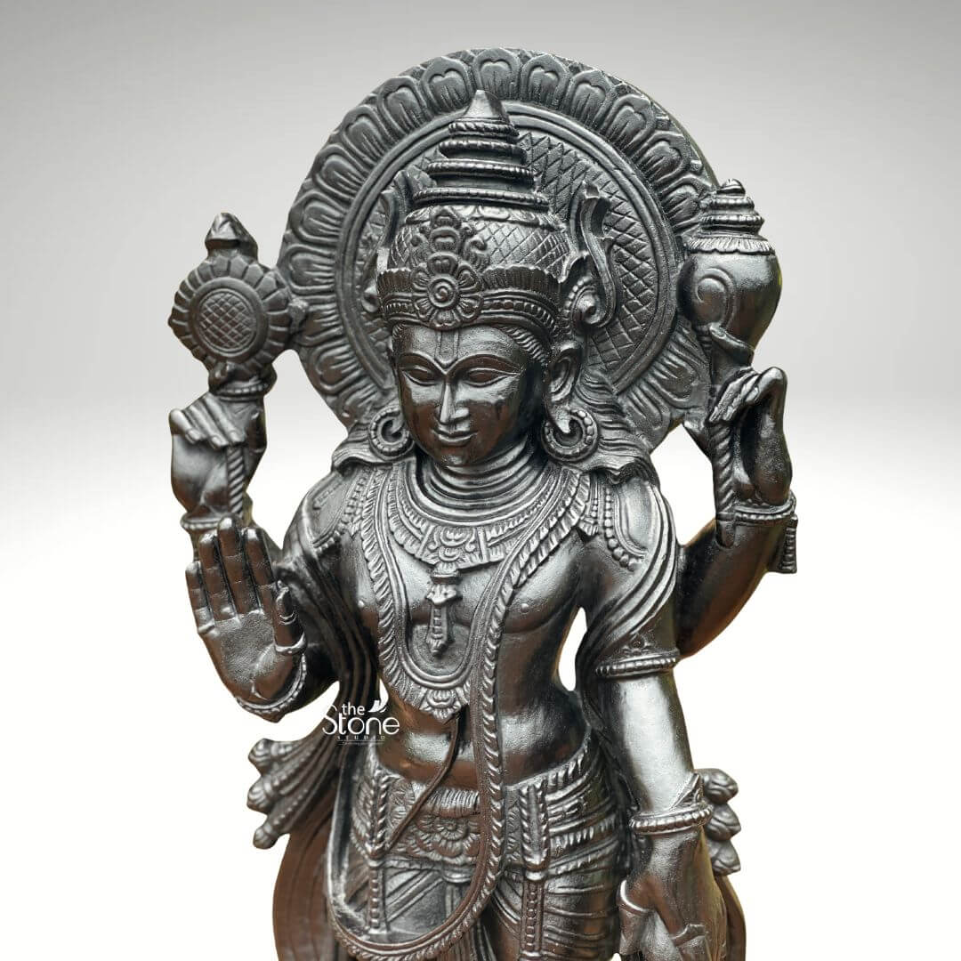 Black Stone Narayana Swamy Statue 3.5ft 4 A detailed close-up photograph captures the upper body and serene face of a majestic forty-one-inch statue of Lord Narayana Swamy meticulously hand-carved from a single block of deep black granite. The deity's face conveys a profound sense of inner peace and divine compassion with half-closed eyes and a gentle benevolent smile. Its head is adorned with an intricately carved towering mukuta featuring detailed patterns and a central floral motif all defined with exceptional precision against the dark stone. The fine natural crystalline structure and rich dark matte-to-satin finish characteristic of high-quality black granite are evident throughout the sculpture. The four-armed form of Lord Narayana is depicted with elaborate shallow-relief carvings of traditional South Indian temple jewelry including multiple necklaces intricate armlets bracelets and a sacred thread all rendered with exceptional detail. The deity's four arms radiate outwards from the central figure showcasing the artisan's skill in defining each attribute. The upper right hand holds the chakra while the upper left hand holds the shankha. The lower right hand is shown in the abhaya mudra a gesture of fearlessness and blessing. A small portion of the gada is visible in the lower left hand completing the traditional iconography. The statue conveys a sense of monumental presence and spiritual majesty with deep relief carvings defining the folds of the garments and the texture of the ornaments against the smooth skin. A grand intricately carved prabhavali forms a halo behind the deity adorned with stylized floral and geometric patterns. The statue is presented against a clean neutral graduated grey background ensuring the focus remains entirely on the detailed figure of Narayana Swamy and its artistic craftsmanship. The black granite material ensures durability and a timeless quality making this large statue a commanding presence for any devotional space.