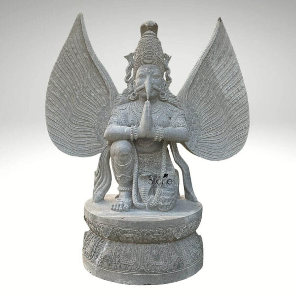 A detailed front-facing photograph captures a majestic 5.5-foot-tall Garuda statue meticulously hand-carved from a single block of natural grey stone. The statue depicts the mythical bird-deity Garuda in a reverent kneeling posture on a robust circular lotus-carved pedestal base. His hands are pressed together in the Anjali Mudra a gesture of prayer and devotion held near his chest. Garuda has a powerful human-like torso with an eagle-like face defined by a prominent beak piercing eyes and a traditional crown with intricate patterns. His body is adorned with finely carved traditional ornaments, including layered necklaces armbands bracelets and a sacred thread all rendered in deep relief that highlights the skill of the artisan. A striking feature of this grey stone Garuda statue is its massive fully spread wings which rise dramatically behind him mirroring his height. Each wing feather is individually and delicately carved creating a texture that is both visually detailed and structurally impressive. The grey stone itself possesses a natural slightly matte finish with subtle crystalline variations, giving the sculpture an authentic ancient feel and underscoring the natural quality of the material. The entire form balances grace with power capturing the essence of the revered celestial mount of Lord Vishnu. This five-and-a-half-foot grey stone Garuda statue is an exceptional example of traditional Indian stone carving, making it a perfect centerpiece for any article discussing Garuda sculptures or Hindu iconography. Its commanding size and detailed craftsmanship make it suitable for prominent placement in a home temple a garden sanctuary or as an architectural element in a larger space. The choice of durable grey stone ensures its longevity making it a timeless investment for spiritual and aesthetic appreciation. The image clearly displays the unique iconographic elements and the high level of artistry that define such a premium stone statue.
