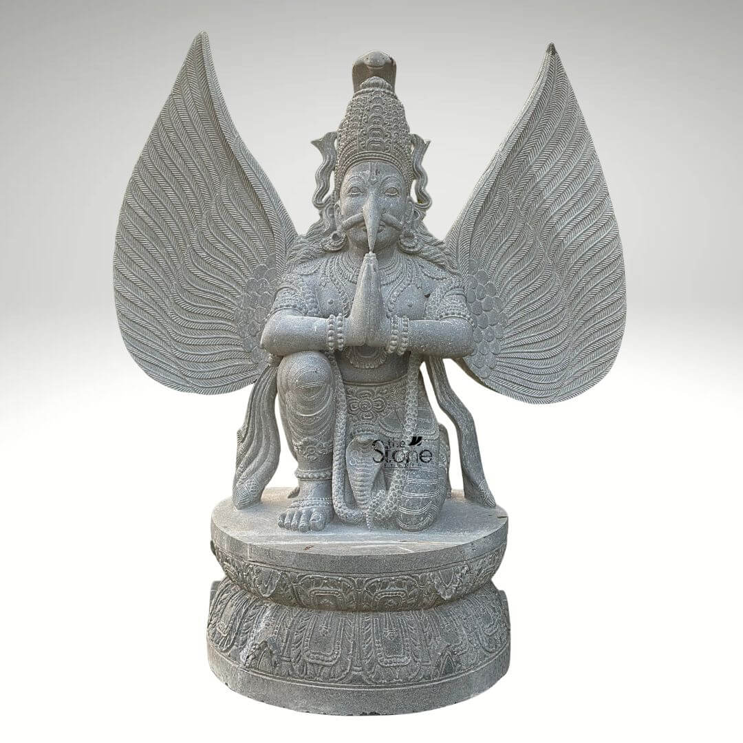 Grey Stone Garuda Statue 5.5ft 1 A detailed front-facing photograph captures a majestic 5.5-foot-tall Garuda statue meticulously hand-carved from a single block of natural grey stone. The statue depicts the mythical bird-deity Garuda in a reverent kneeling posture on a robust circular lotus-carved pedestal base. His hands are pressed together in the Anjali Mudra a gesture of prayer and devotion held near his chest. Garuda has a powerful human-like torso with an eagle-like face defined by a prominent beak piercing eyes and a traditional crown with intricate patterns. His body is adorned with finely carved traditional ornaments, including layered necklaces armbands bracelets and a sacred thread all rendered in deep relief that highlights the skill of the artisan. A striking feature of this grey stone Garuda statue is its massive fully spread wings which rise dramatically behind him mirroring his height. Each wing feather is individually and delicately carved creating a texture that is both visually detailed and structurally impressive. The grey stone itself possesses a natural slightly matte finish with subtle crystalline variations, giving the sculpture an authentic ancient feel and underscoring the natural quality of the material. The entire form balances grace with power capturing the essence of the revered celestial mount of Lord Vishnu. This five-and-a-half-foot grey stone Garuda statue is an exceptional example of traditional Indian stone carving, making it a perfect centerpiece for any article discussing Garuda sculptures or Hindu iconography. Its commanding size and detailed craftsmanship make it suitable for prominent placement in a home temple a garden sanctuary or as an architectural element in a larger space. The choice of durable grey stone ensures its longevity making it a timeless investment for spiritual and aesthetic appreciation. The image clearly displays the unique iconographic elements and the high level of artistry that define such a premium stone statue.