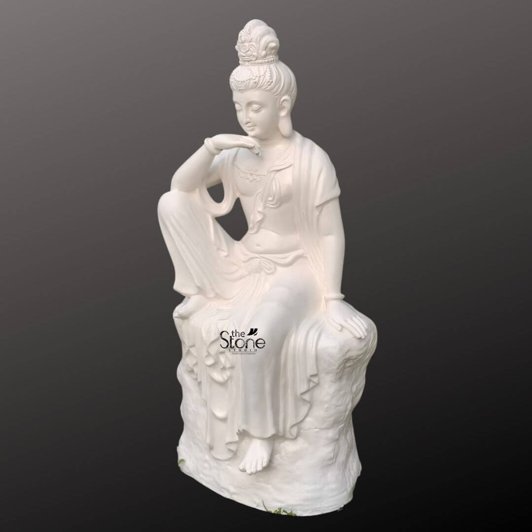 Seated Kuan Yin Statue 3ft 2 A 3 feet fibre Guanyin meditation statue is presented in a clean studio-style image against a soft dark grey gradient background allowing the sculpture to stand out clearly as the central focus. The statue depicts Guanyin also known as Kuan Yin in a graceful seated pose on a textured rock-like pedestal. One leg is bent upward with the elbow resting lightly on the knee while the hand rises gently near the chin creating a thoughtful meditative posture. The other hand rests softly on the seat beside the body and one foot extends downward giving the statue a relaxed yet elegant balance. The face is turned slightly downward with closed or lowered eyes delicate features and a calm expression that conveys compassion inner peace and spiritual reflection. The white fibre Kuan Yin statue is richly sculpted with flowing robes that drape naturally over the body and cascade in soft folds across the stone-like base. The figure wears a detailed ornamental crown or headdress along with subtle jewellery accents around the neck and wrists that add to the devotional beauty of the piece. The smooth white finish enhances the serene quality of the sculpture while the carved textures of the pedestal create contrast and visual depth. The side angle of the image helps reveal the dimensional craftsmanship of the Guanyin meditation statue highlighting the curve of the posture the layered robes and the elegant contours that make the piece both artistic and spiritually expressive. This image connects strongly to an article about a Guanyin Meditation Statue by clearly showing the peaceful form material and decorative appeal of a 3 feet fibre Kuan Yin statue. It presents the sculpture as a meaningful choice for meditation rooms spiritual corners peaceful indoor décor temple settings wellness spaces or tranquil garden-inspired interiors. The overall visual impression emphasizes Guanyin as a symbol of mercy compassion, and mindfulness while also showing how this seated fibre statue can serve as a refined decorative focal point. Its serene pose graceful styling and devotional presence make it an ideal representation of a Guanyin meditation statue for those seeking calm spiritual beauty and elegant sacred décor.