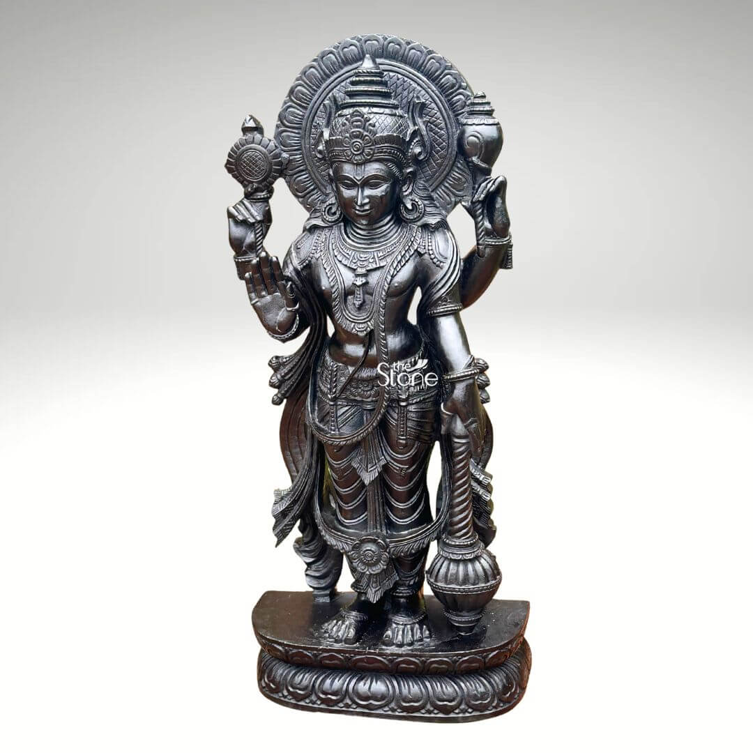 Black Stone Narayana Swamy Statue 3.5ft 5 A detailed full-body photograph captures a majestic forty-one-inch-tall statue of Lord Narayana Swamy meticulously hand-carved from a single solid block of deep black stone. The deity is presented in a traditional standing posture upon an intricately carved circular lotus pedestal which features detailed petal layers and a decorative base. Narayana is depicted with four arms radiating divine grace and power making it an ideal sacred art piece for a home temple or place of worship. The statue exhibits a rich dark matte-to-satin finish characteristic of high-quality black stone which absorbs light and highlights the depth and complexity of the carvings. The four-armed form of Lord Narayana holds traditional symbolic attributes each intricately defined against the dark stone. His upper right hand holds the chakra while his upper left hand holds the shankha. His lower right hand is shown in the abhaya mudra a gesture of fearlessness and blessing and his lower left hand rests gracefully holding the gada. The deity is adorned with elaborate shallow-relief carvings of traditional South Indian temple jewelry including multiple necklaces armlet bracelets a sacred thread and a decorative mukuta all rendered with exceptional precision. The statue conveys a sense of profound spiritual calm and majesty with the intricate details of his flowing dhoti with elaborate folds the detailed garlands and the serene facial expression all contributing to its devotional appeal. The black stone material ensures durability and a timeless quality making this large statue a commanding presence. The statue is presented against a clean neutral graduated grey background ensuring the focus remains entirely on the detailed figure of Narayana Swamy and its artistic craftsmanshi perfectly illustrating its suitability as a devotional focal point.