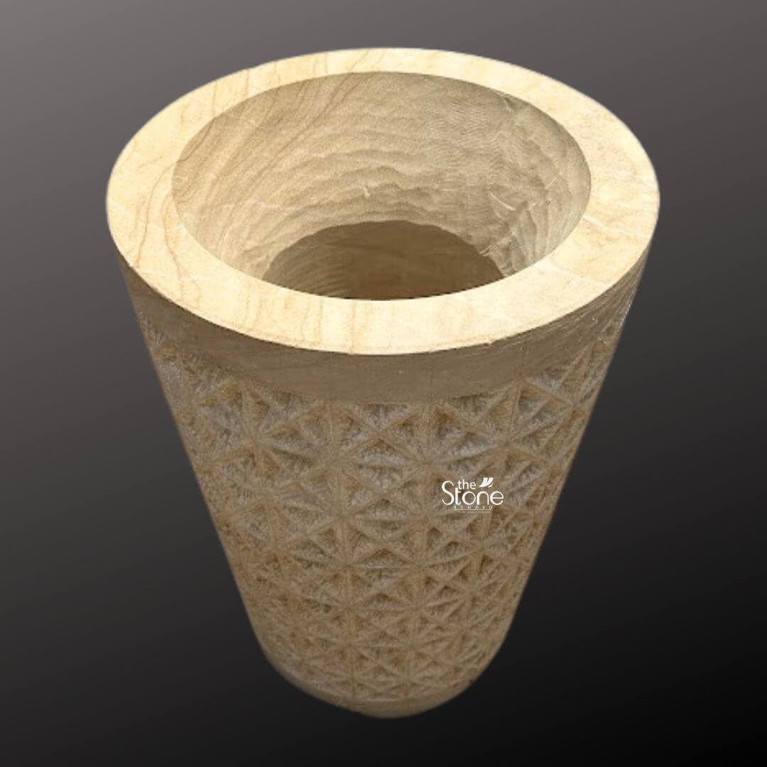 Natural Sandstone Planter Pot 2ft 5 A high-angle studio photograph of a magnificent 2-foot tall hand-carved stone planter pot presented against a dark neutral gray gradient background. The planter is crafted from a solid block of natural warm-toned sandstone exhibiting a rich earthy texture with subtle color variations that highlight its geological origin. It is fashioned into a classic slightly tapered cylindrical form providing a robust and stable base. The interior is deeply hollowed revealing the raw rugged texture of the stone resulting from the carving process while the thick smooth uncarved rim offers a clean contrast. The central body of the stone planter pot is adorned with an intricate deeply incised geometric pattern. This pattern consists of a repeating tessellation of diamond and triangular facets, creating a textured three-dimensional surface that catches the light and adds significant visual depth. The precision and detail of the carving demonstrate a high level of artisanal skill transforming the functional object into a piece of sculptural art. The natural porosity of the sandstone is clearly visible emphasizing its suitability for outdoor garden environments and its ability to breathe which is beneficial for plant health. This substantial hand-carved stone planter standing at 2 feet is an ideal choice for showcasing large houseplants indoors or as a striking focal point in a garden patio or entryway. Its natural stone construction and timeless geometric design make it a versatile piece that complements various decor styles from rustic to modern. The image clearly conveys the planter's scale texture and elaborate design making it a perfect visual reference for an article discussing hand-carved stone garden elements and artisanal craftsmanship.