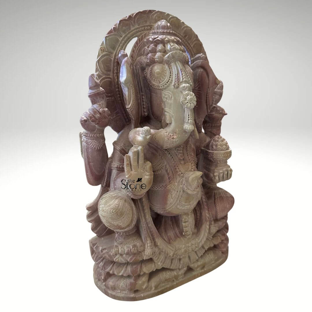 Pink Stone Ganpati Idol for Home 12" 2 A three-quarter view photograph showcases a meticulously handcrafted twelve-inch Lord Ganesha statue sculpted from a single piece of natural pink stone. The statue depicts the elephant-headed Hindu deity in a seated meditative pose with one leg gently folded over the other resting on a multi-tiered lotus-carved pedestal base. The pink stone exhibits a soft variegated hue ranging from pale rose to deeper mauve with subtle natural veining and crystalline inclusions throughout giving the idol a warm and earthy texture. The sculpture is ighly detailed with intricate carvings covering every aspect of the divine form. Ganesha is adorned with traditional deeply carved jewelry including elaborate layered necklaces armbands anklets and a richly patterned crown that frames his serene face. His eyes are half-closed in contemplation, and his trunk curves gracefully to his left holding a sweet modak. The four-armed deity holds symbolic attributes in his hands, all rendered with fine precision: his upper right hand holds a goad his upper left hand holds a noose his lower right hand is raised in the Abhaya mudra and his lower left hand holds a bowl overflowing with modaks. A decorative prabhavali with intricate floral and geometric motifs rises behind his head completing the divine iconography. This twelve-inch pink stone Ganesha statue is an exemplary piece of Indian stone carving art making it a perfect subject for an article on handcrafted Lord Ganesha statues. The compact size suggests its suitability for a home altar a prominent shelf or as a decorative centerpiece bringing a sense of spiritual tranquility and cultural artistry to any interior space. The choice of pink stone adds a gentle inviting color to the decor. The detailed craftsmanship and traditional iconography showcased in this image highlight the enduring cultural and religious significance of Ganesha in Indian households. The photograph's clear focus on the statue's texture form and craftsmanship provides a comprehensive view for the viewer.