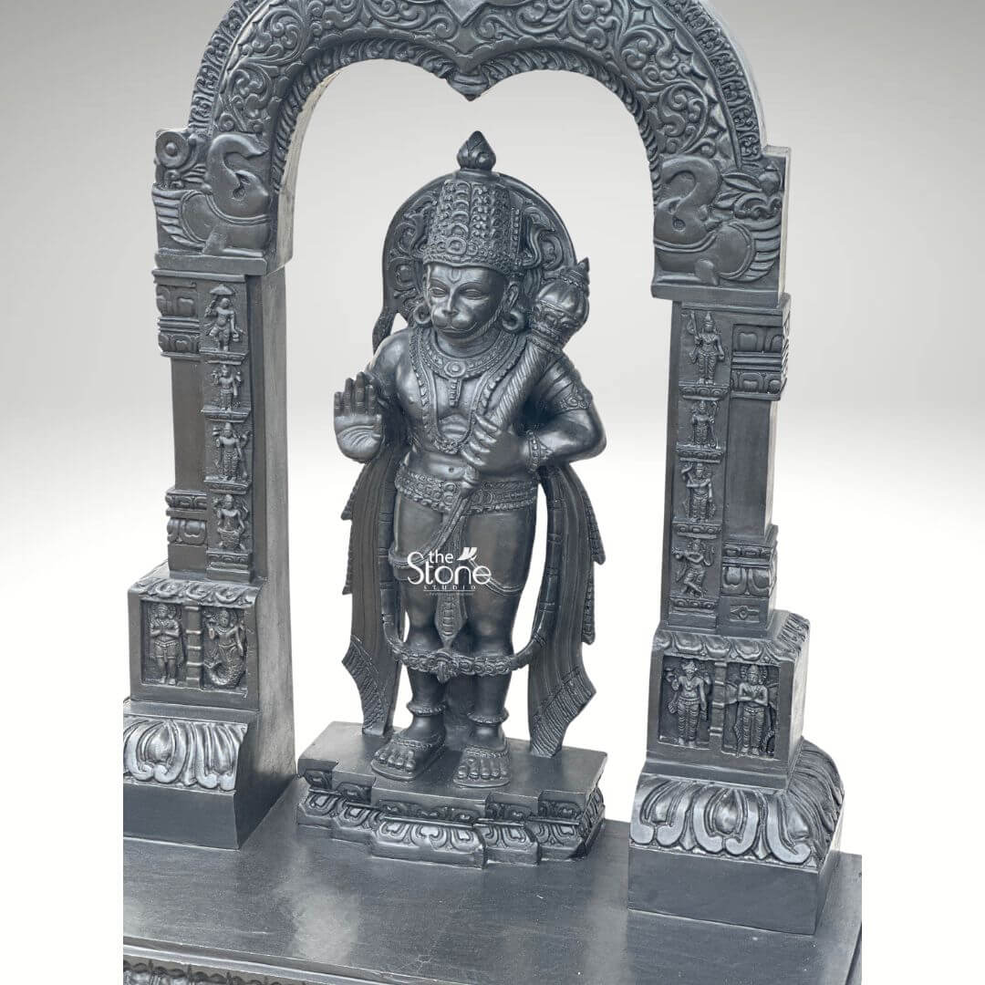 Black Stone Hanuman Statue with Arch 3ft 2 A close-up view reveals a magnificent 3-foot Handcrafted Black Stone Hanuman Murti intricately carved and presented within an ornate black stone arch against a crisp white background. Lord Hanuman is depicted in a standing posture exuding strength devotion and readiness for service. His form is richly detailed with an elaborate crown traditional jewelry adorning his neck, chest and arms and finely sculpted garments that suggest flowing fabric. In his left hand he steadfastly holds his gada a potent symbol of his immense power and valor while his right hand is raised in a gesture of Abhaya Mudra offering fearlessness and protection. The craftsmanship of this Black Stone Hanuman statue is exceptional with every detail meticulously brought to life in the deep lustrous black stone. The facial features including his serene yet determined expression and the subtle rendering of his monkey-god characteristics are precise. The musculature of his body is subtly suggested conveying his legendary strength. He stands on a multi-tiered pedestal also carved from black stone which is integrated into the larger base of the archway. The statue is dramatically framed by a beautifully carved arch that features intricate traditional patterns floral motif and smaller divine figures or mythological scenes on its pillars and archway characteristic of ancient temple artistry. These surrounding elements enhance the spiritual grandeur and cultural richness of the entire piece. This 3-foot Handcrafted Black Stone Hanuman Murti with its powerful presence and exquisite detailing within the arched framework serves as an ideal representation for an article focused on such a divine sculpture symbolizing unwavering loyalty courage and spiritual strength.