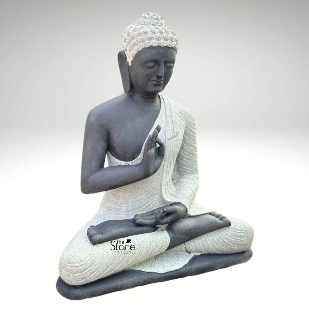 Two Tone Buddha Stone Statue for Home 3ft 5 A serene hand-carved Blackstone Buddha statue sits in a traditional meditative pose presented in a clean studio setting against a soft grey background. The three-foot-tall statue showcases a striking two-tone finish with the Buddha's body and head carved from a deep dark blackstone exhibiting a smooth polished texture that highlights the rich dark tones. This contrasts beautifully with his monastic robes which are rendered in a contrasting lighter grey or off-white stone featuring a subtle granular texture. This distinct separation emphasizes the form of the robes as they drape over his crossed legs and shoulders adding depth and visual interest to the sacred art. The Buddha is depicted seated with his legs crossed in a full lotus position on a simple integrated dark blackstone base. His hands are gracefully positioned in the vitarka mudra symbolizing discussion and the transmission of teachings with the right hand raised and the thumb and index finger forming a circle while the left hand rests in his lap. His facial expression is calm and contemplative with downcast eyes a gentle smile and tightly curled hair leading to a cranial ushnisha all rendered with precise craftsmanship that honors traditional iconography. The overall composition evokes a sense of peace tranquility and spiritual harmony. This three-foot Blackstone Buddha statue is an ideal illustration for an article discussing Handcrafted Buddha Sculpture demonstrating how such a piece can transform an indoor or outdoor space into a peaceful sanctuary. The unique dual-color and dual-texture stone work makes it a stunning focal point for a living room entryway or dedicated meditation area while its natural stone texture also harmonizes beautifully with surrounding plant life in a garden setting. The traditional design brings a sense of sacred history and calm to any home interior or exterior while the durable blackstone construction ensures it is both a beautiful and a meaningful addition to the home.