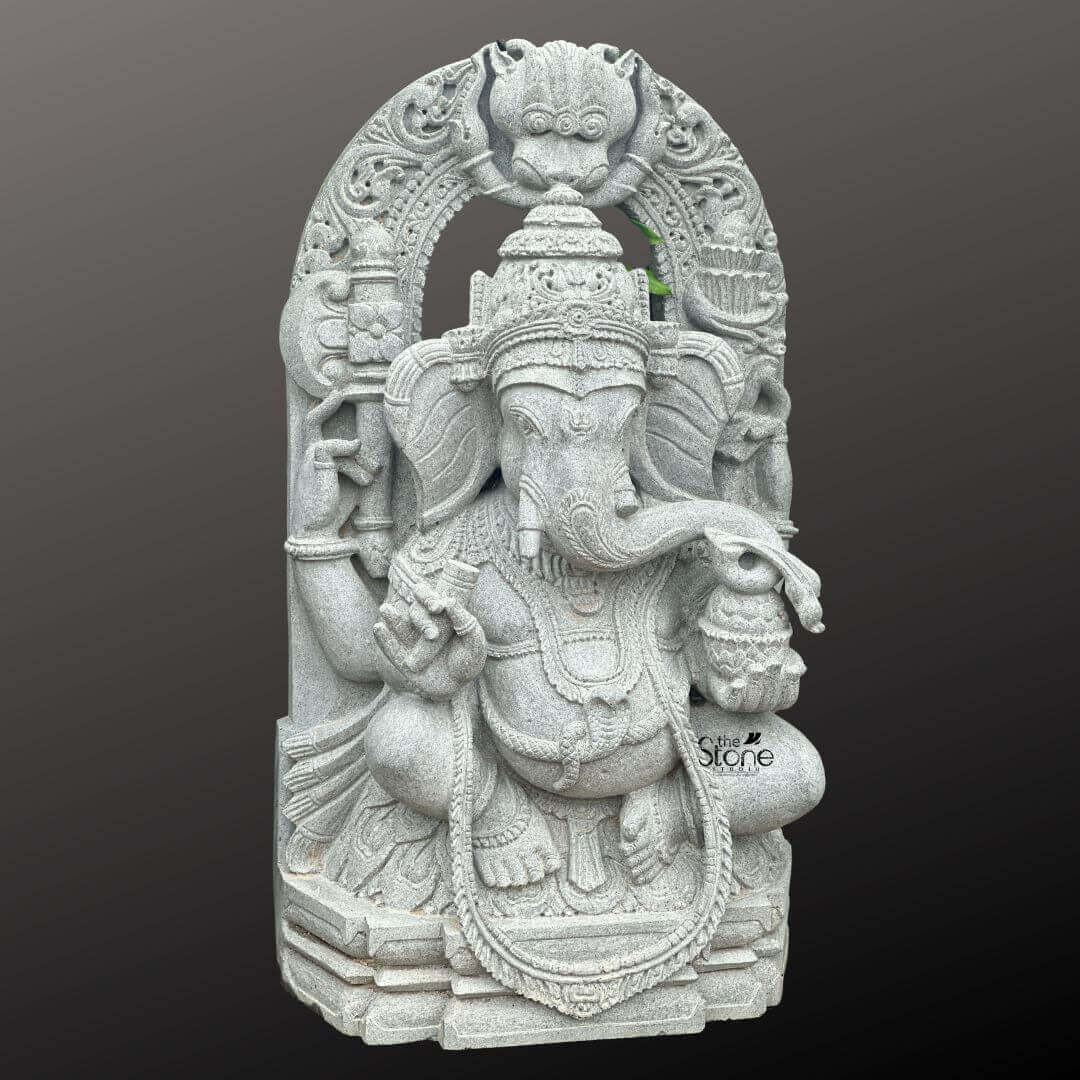Ganesha Statue with Makara Torana 3ft 4 A magnificent 3-foot Handcrafted Ganesha Stone Idol intricately carved from what appears to be a light grey stone stands powerfully against a dark gradient background. The statue depicts Lord Ganesha in a regal seated posture exuding benevolence wisdom and an aura of divine grace. His iconic elephant head is finely sculpted complete with large ears expressive eyes and his distinctive broken tusk. He is adorned with an elaborate crown multiple layers of traditional jewelry including necklaces armlets and a sacred thread draped across his chest all rendered with exceptional detail in the light grey stone emphasizing the handcrafted quality. Ganesha is portrayed with four arms holding his customary attributes a modak in one hand an axe or goad in another and his lower right hand extended in a gesture of blessing offering reassurance and protection. The intricate details on his garments the patterns on his halo and the careful sculpting of his prominent pot belly are all clearly visible showcasing the high artistry of the carving. The light grey stone emphasizes these textures and details lending a serene and timeless elegance to the deity and suggesting a weathered ancient feel that speaks to its artisanal creation. The statue is dramatically framed by a grand ornate arch that features exquisite mythical creature motifs and intricate traditional carvings along its entire span and supporting pillars. The top of the arch features a Kirtimukha a fierce lion-like face a common protective motif. This entire elaborate structure rests on a multi-tiered base also carved from the same light grey stone providing stability and enhancing its grand temple-like presence. This 3-foot Handcrafted Ganesha Stone Idol with its impressive arch is an exquisite example of religious artistry designed to bring blessings peace and aesthetic grandeur to any space perfectly aligning with an article focusing on such a revered Ganpati sculpture.