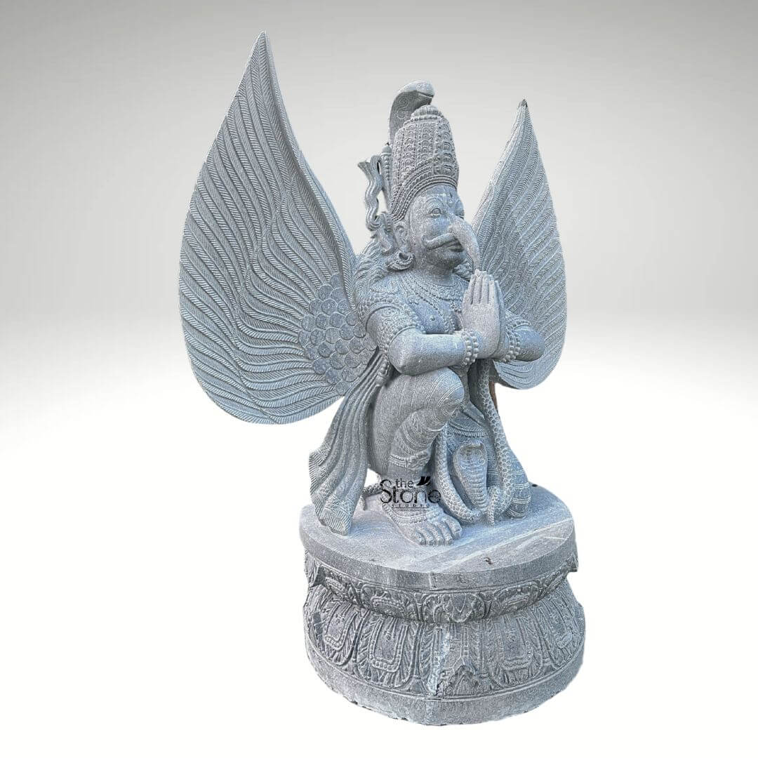 Grey Stone Garuda Statue 5.5ft 5 A three-quarter view photograph captures a majestic 5.5-foot-tall Garuda statue meticulously hand-carved from a single block of natural grey stone. The statue presents the mythical bird-deity Garuda in a reverent kneeling posture on a robust circular lotus-carved pedestal base. His hands are pressed together in the Anjali Mudra, a gesture of prayer and devotion, held near his chest. Garuda has a powerful human-like torso with an eagle-like face defined by a prominent beak piercing eyes and a traditional crown with intricate patterns. His body is adorned with finely carved traditional ornaments including layered necklaces armbands bracelets and a sacred thread all rendered in deep relief that highlights the skill of the artisan. A striking feature of this grey stone Garuda sculpture is its massive fully spread wings which rise dramatically behind him mirroring his height. Each feather is individually and delicately carved creating a texture that is both visually detailed and structurally impressive. The grey stone itself possesses a natural slightly matte finish with subtle crystalline variations, giving the sculpture an authentic ancient feel and underscoring the natural quality of the material. The carving exhibits a high level of detail from the individual feather points to the segmented body of a serpent coiled at his feet reflecting the skill and patience of the master artisan who created it. This five-and-a-half-foot handcrafted Garuda sculpture is an exceptional example of traditional Indian stone carving making it a perfect centerpiece for any article discussing Garuda sculptures or Hindu iconography. Its commanding size and detailed craftsmanship make it suitable for prominent placement in a home temple a garden sanctuary or as an architectural element in a larger space. The choice of durable grey stone ensures its longevity making it a timeless investment for spiritual and aesthetic appreciation. The image clearly displays the unique iconographic elements and the high level of artistry that define such a premium stone statue.