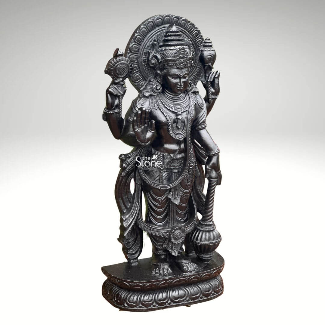Black Stone Narayana Swamy Statue 3.5ft 6 A detailed full-body photograph captures a majestic forty-one-inch-tall statue of Lord Narayana Swamy meticulously hand-carved from a single solid block of deep black stone. The deity is presented in a traditional standing posture upon an intricately carved circular lotus pedestal which features detailed petal layers and a decorative base. Narayana is depicted with four arms radiating divine grace and power making it an ideal sacred art piece for a home temple or place of worship. The statue exhibits a rich dark matte-to-satin finish characteristic of high-quality black stone which absorbs light and highlights the depth and complexity of the carvings. The four-armed form of Lord Narayana holds traditional symbolic attributes each intricately defined against the dark stone. His upper right hand holds the chakra while his upper left hand holds the shankha. His lower right hand is shown in the abhaya mudra a gesture of fearlessness and blessing and his lower left hand rests gracefully holding the gada. The deity is adorned with elaborate shallow-relief carvings of traditional South Indian temple jewelry including multiple necklaces armlet bracelets a sacred thread and a decorative mukuta all rendered with exceptional precision. The statue conveys a sense of profound spiritual calm and majesty with the intricate details of his flowing dhoti with elaborate folds the detailed garlands and the serene facial expression all contributing to its devotional appeal. The black stone material ensures durability and a timeless quality making this large statue a commanding presence. The statue is presented against a clean neutral graduated grey background ensuring the focus remains entirely on the detailed figure of Narayana Swamy and its artistic craftsmanship perfectly illustrating its suitability as a devotional focal point.