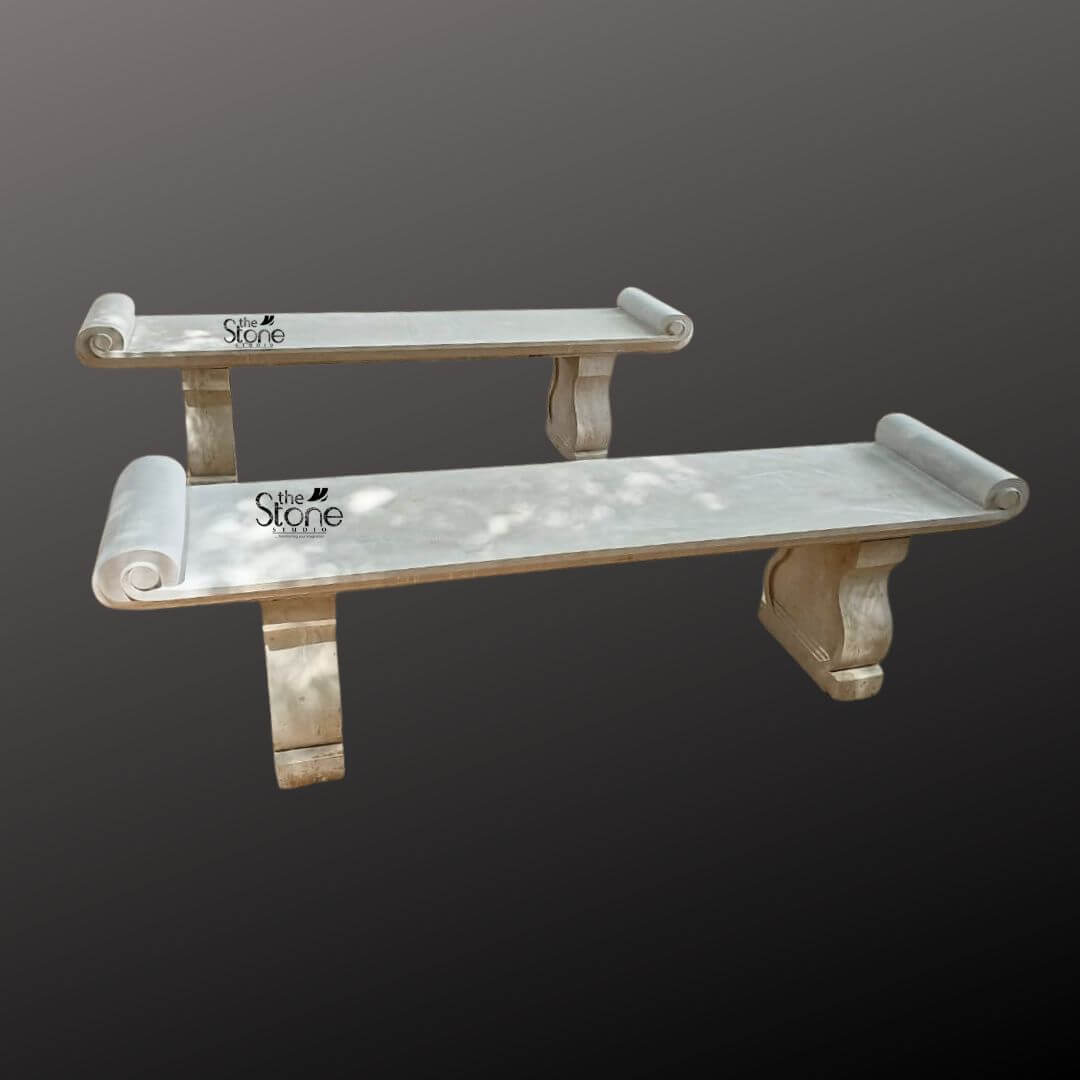 Sandstone Garden Bench 6ft 2 A stunning hand-carved six-foot sandstone bench stands as the focal point against a dark gradient studio background. The bench is meticulously crafted from natural light beige sandstone showcasing a beautifully textured porous surface that adds an authentic rustic charm. Its long flat seat is supported by two substantial intricately sculpted legs. These legs feature a graceful S-curve profile resting on sturdy block-like bases that anchor the entire piece. The craftsmanship is evident in the smooth hand-hewn finish of the stone which catches the light and highlights its natural variations in color and texture from pale cream to subtle warm brown tones. The design of this handcrafted bench is distinctly classical inspired by historical European garden furniture. The seat itself is not plain it terminates at both the left and right ends with large perfectly formed scrolled volutes or rolled ends. These sculptural elements not only provide comfortable armrests but also define the bench's aesthetic lending it a sophisticated and timeless appeal. The overall form is balanced and substantial suggesting durability and a permanent place within a garden landscape. The stone appears heavy and solid capable of withstanding the elements for decades. This six-foot sandstone bench is the perfect embodiment of a functional and decorative Handcrafted Stone Garden Bench. Its generous length provides ample seating for multiple people while its robust stone construction makes it an ideal choice for outdoor settings like patios courtyards or nestled within garden pathways. The light neutral color of the sandstone harmonizes beautifully with surrounding greenery flowers and other natural elements. This bench is not just a piece of furniture it is a sculptural addition to a garden designed to offer a comfortable and contemplative spot to rest and enjoy the beauty of the outdoors.