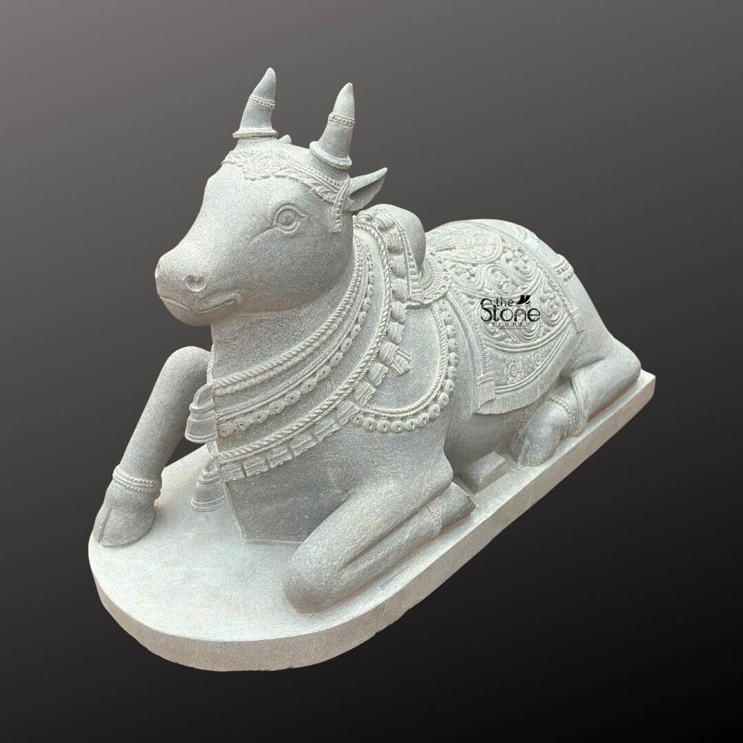Nandikeshwara Stone Statue 45" 4 A three-quarter front view photograph captures a magnificent hand-carved stone statue of Nandi the divine bull and mount of Lord Shiva. The statue is sculpted from a single solid block of dark grey blackstone which has been polished to a deep matte finish emphasizing the rich deep color and intricate sculptural details. Nandi is depicted in a traditional serene reclining posture resting upon a simple elevated rectangular blackstone plinth. His head is alert and turned slightly to his left gazing with a calm benevolent expression. The fine-grained texture of the stone is visible giving the piece an authentic ancient feel. Nandi is richly adorned with traditional deeply carved ornaments that showcase the skill of the artisan. He wears multiple layered necklaces of varying designs including beaded chains and bell-shaped pendants along with a decorative forehead band and ear ornaments. A large ornate blanket with elaborate floral paisley and geometric patterns in high relief covers his back and hindquarters extending down the sides with fringed borders. The carving extends to his legs which are decorated with anklets and his horns and humps are carefully rendered. The overall form is powerful yet graceful capturing the spiritual and aesthetic essence of Nandi. This Handcrafted Blackstone Nandi Statue is an exceptional example of traditional South Indian temple carving art making it a perfect subject for an article discussing such sacred sculptures. Its size detailed craftsmanship and traditional iconography make it suitable for placement in a home shrine a temple garden or as a central exhibit capturing the spiritual significance and cultural heritage of the deity. The durable blackstone material ensures the statue's longevity making it a timeless investment for both spiritual devotion and artistic appreciation. The photograph's clear focus on the statue's texture form and ornamentation provides a comprehensive view for the viewer highlighting the unique characteristics of blackstone carving.