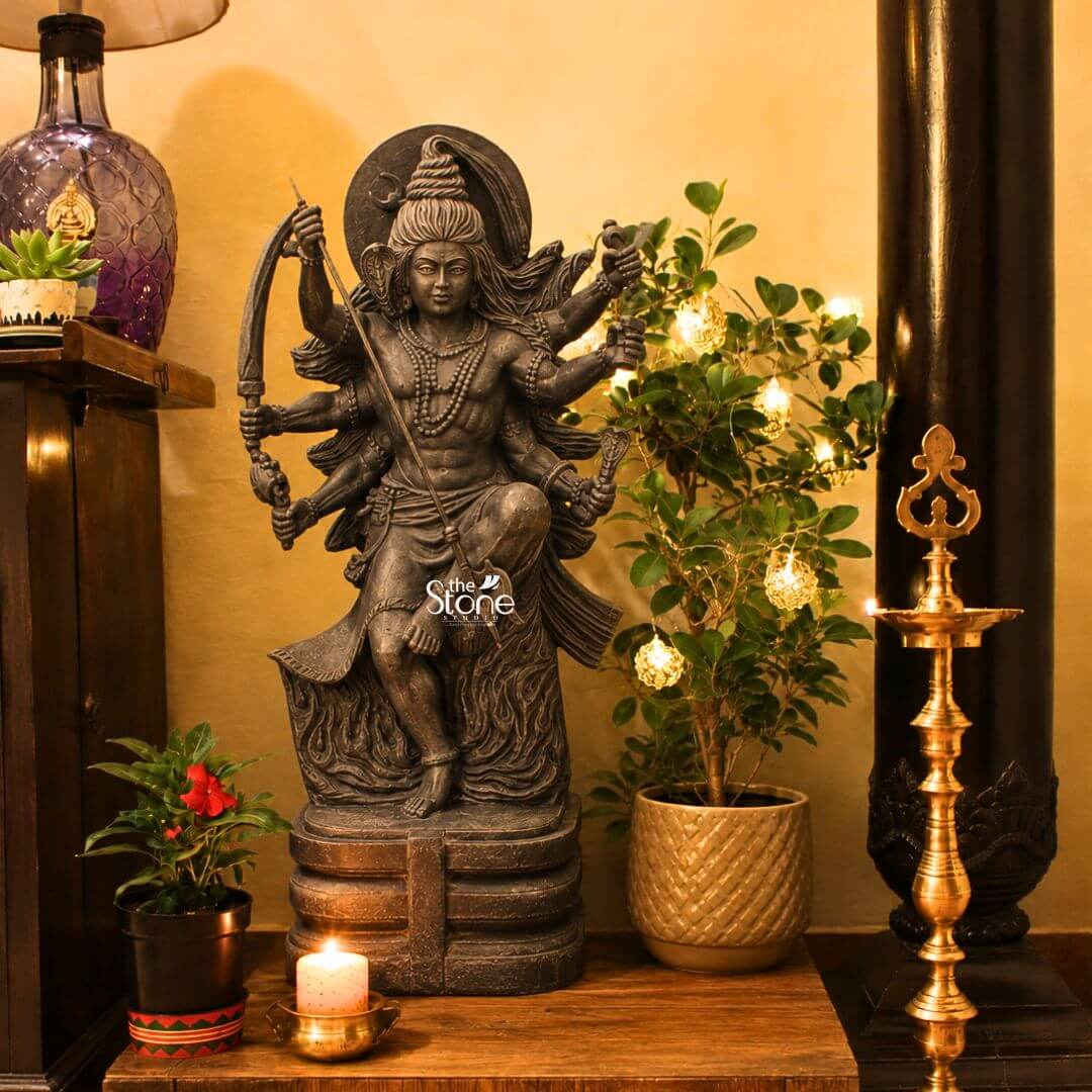 2 Feet Tandava Shiva Statue 3 A beautifully composed photograph captures a handcrafted Tandava Shiva statue standing approximately 2 feet tall as the central deity in a home altar. The statue hand-carved from a single block of dense polished black stone depicts Lord Shiva in the dynamic and powerful Ananda Tandava pose the dance of cosmic bliss set upon a detailed multi-tiered lotus pedestal. Shiva is portrayed with ten arms fanned out to convey movement holding various divine attributes including a trident and a damaru drum. His matted hair is piled high adorned with a crescent moon and a serpent and flares out dramatically emphasizing the energy and rhythm of his celestial dance. The black stone used for the statue has a deep rich hue with a subtle, waxy luster, giving it a powerful and reverent presence that complements the surrounding decor. The intricate details of the carving, from the delicate folds of Shiva's garment to the fierce yet serene expression on his face and the form of the demon Muyalakan subdued beneath his foot showcase the exquisite craftsmanship involved in creating this handcrafted piece. The statue is placed on a rustic wooden console table against a warm textured golden-yellow wall flanked by potted plants including a leafy ficus adorned with small warm fairy lights and a traditional ornate brass oil lamp standing tall to the right. This 2-foot black stone Tandava Shiva statue stands as a perfect embodiment of Indian temple art and a meaningful centerpiece for any sacred space. Its compact size allows for placement on an altar or a display table where its intricate details and spiritual energy can be admired. The dark monolithic stone gives it a timeless quality while the powerful iconography of the Tandava pose evokes a sense of cosmic rhythm creation and destruction making it an ideal choice for anyone seeking to bring a touch of Eastern spirituality and artistic heritage into their home. The photograph presents the statue within its decorative context bathed in the warm soft light of the lamp and fairy lights enhancing its divine aura.