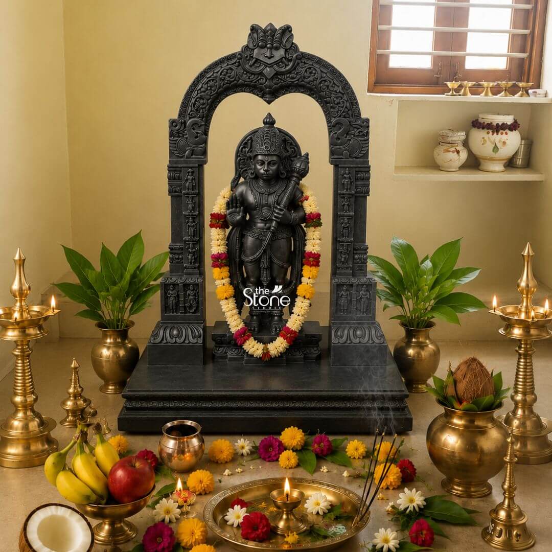 Black Stone Hanuman Statue with Arch 3ft 3 A beautifully adorned 3-foot Blackstone Hanuman Murti with Makara Torana is the focal point of a vibrant puja setup set against a warm light yellow wall in a traditional home temple. Lord Hanuman is depicted standing radiating strength and devotion intricately carved from deep black stone. He is garlanded with a long colorful mala of yellow white and red flowers which elegantly drapes over his form. In his left hand he firmly holds his gada a symbol of his immense power while his right hand is raised in a gesture of blessing. The statue is dramatically framed by a grand black stone archway known as a Makara Torana which features exquisite mythical creature motifs at its base and intricate traditional carvings along its entire span and pillars. The detailing on the black stone from Hanuman's crown and jewelry to the ornate patterns on the torana is exceptionally precise showcasing the artistry of the sculpture. The entire structure rests on a multi-tiered black stone base lending a majestic and stable presence. The devotional space around Hanuman is richly decorated with traditional items. Two tall gleaming brass oil lamps with multiple wicks are lit casting a warm glow on either side. In front a brass tray holds a lit diya incense sticks emitting smoke and loose flowers of various colors. Bowls of fresh fruits like bananas and apples a split coconut and brass pots with green plants further enhance the sacred atmosphere. This elaborate arrangement with the 3-foot Blackstone Hanuman statue and its impressive Makara Torana creates a deeply spiritual and aesthetically rich home temple perfectly aligning with an article highlighting this specific type of Hanuman Murti.