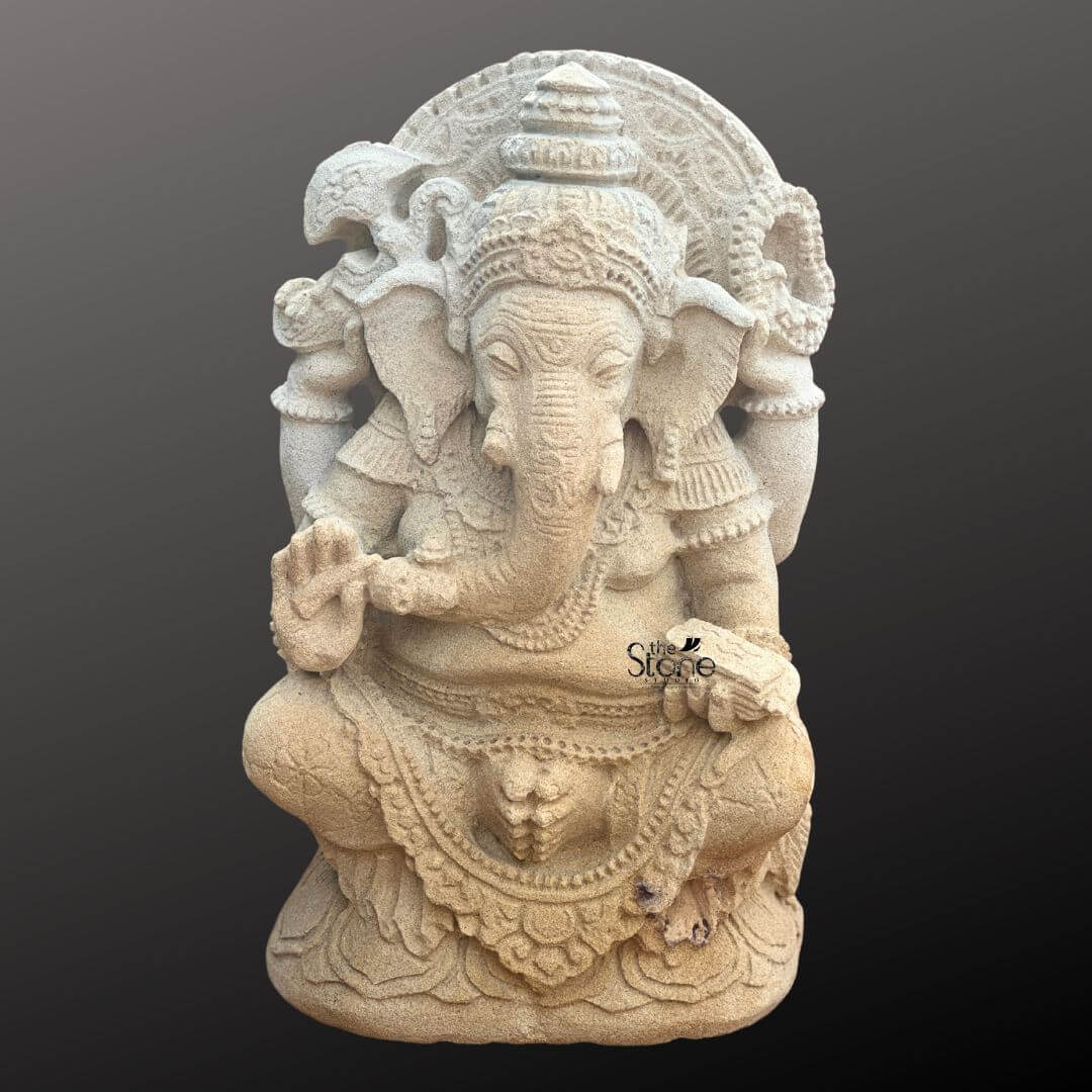 Indonesian Ganesha Idol for Home 2ft 1 A full-body studio photograph of a magnificent 2-foot tall Indonesian Ganesha Idol meticulously hand-carved from warm beige sandstone presented against a dark neutral gray gradient background that accentuates its rich texture. The statue depicts the elephant-headed deity Lord Ganesha in a traditional seated posture upon a detailed lotus pedestal embodying profound tranquility and wisdom. The craftsmanship is highly detailed showcasing traditional Balinese artistic influence with ornate carvings on his headdress jewelry and flowing garments. Ganesha is depicted with four arms holding symbolic attributes that convey his divine powers. His upper right hand holds an axe and his upper left hand holds a broken tusk while his lower right hand is raised in a gesture of blessing and his lower left hand holds a sweet that his trunk is tasting. The natural porosity and tonal variations of the sandstone are visible adding an earthy elegance and a sense of age to the piece. The statue's serene expression and detailed ornamentation make it a striking example of Indonesian religious art. This 2-foot sandstone Ganesha statue is an ideal centerpiece for a home shrine meditation room or as a decorative element in a living space seeking a touch of Eastern spirituality and cultural heritage. Its compact size allows for prominent placement on an altar a console table or within a garden niche bringing a sense of auspiciousness and divine grace to its surroundings. The image clearly conveys the scale texture and intricate design of the idol making it a perfect visual reference for an article discussing Indonesian Ganesha idols for home decor.