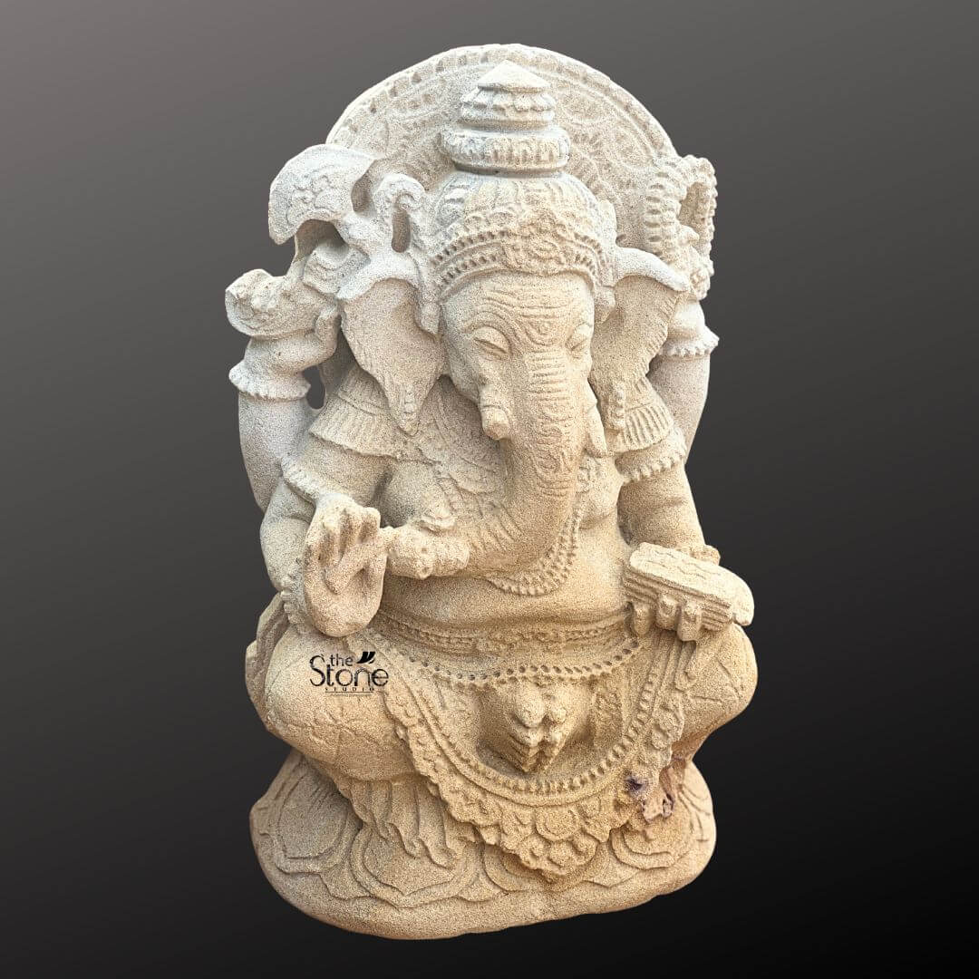 Indonesian Ganesha Idol for Home 2ft 4 A detailed three-quarter view photograph of a traditional Indonesian-style Ganesha statue hand-carved from light-beige sandstone. The statue stands approximately two feet tall and exhibits an antique weathered finish giving it a rich aged patina. The elephant-headed deity is depicted in a seated meditative Lalitasana pose with his right leg folded and his left leg hanging down resting gracefully on a detailed multi-tiered lotus pedestal base. Ganesha is adorned with intricate deeply carved jewelry characteristic of Indonesian artistry including elaborate necklaces armbands anklets and a richly detailed crown with floral and flame motifs. His multiple arms hold symbolic attributes: his upper right hand holds an axe his upper left hand holds a broken tusk and his lower left hand holds a bowl of modak sweets towards which his trunk gently curls. His lower right hand is raised in the Abhaya mudra a gesture of blessing and protection.The sandstone material displays natural texture and color variations enhancing the antique look and the depth of the carving. The craftsmanship reflects the distinct Indonesian stone carvin g tradition with ornate patterns on his garments and the decorative halo framing his head. The figure has a serene expression with eyes half-closed in contemplation. The overall form is robust yet harmoniouscapturing the spiritual grace associated with Indonesian Ganesha iconography. The statue's pose and elaborate adornments are faithful representations of classical Javanese and Balinese sculptural styles, making it an excellent example for the article. This antique-look sandstone sculpture serves as a perfect illustration for an article on Indonesian Style Ganesha showcasing the unique regional characteristics of the deity's representation. The specific iconography includ ing the elaborate crown the type of jewelry and the inclusion of the broken tusk and axe are all hallmarks of Indonesian Hindu art. The weathered finish further connects the piece to the historical and cultural legacy of stone temple carving in Indonesia. The statue is an ideal piece for discussing the evolution and regional variations of Ganesha imagery within the archipelago.