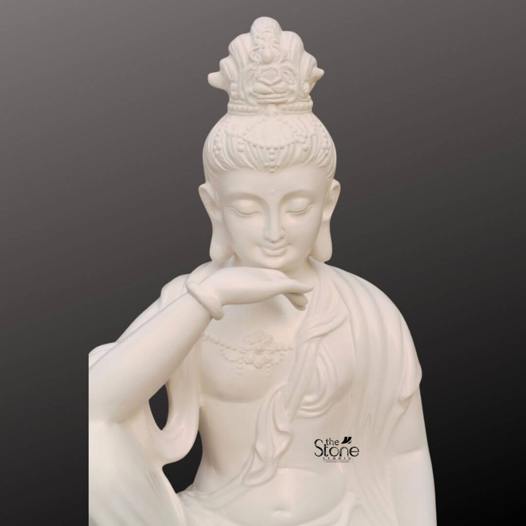 Seated Kuan Yin Statue 3ft 4 A close-up view of a 3 feet fibre Kuan Yin statue shows the upper portion of the sculpture against a smooth dark grey background creating a clean and elegant presentation. The statue depicts Kuan Yin also known as Guanyin with a serene face softly closed eyes a gentle smile and elongated earlobes that enhance the calm and compassionate expression. Her right arm is raised gracefully with the hand resting near the chin in a thoughtful meditative gesture while the draped robe flows softly across the shoulder and chest. The white fibre Kuan Yin statue features delicate ornamental detailing including an intricate crown or headdress above the head and subtle necklace-like embellishment across the chest. The smooth finish of the material gives the sculpture a refined and peaceful appearance while the sculpted folds of the robe add depth and softness to the design. This cropped composition focuses attention on the facial features hand gesture and upper body highlighting the devotional beauty and artistic craftsmanship of the statue. This image connects naturally to an article about a Kuan Yin Statue for Home Décor by showing how a 3 feet Kuan Yin fibre statue can serve as a graceful spiritual accent in interior spaces. The close-up presentation emphasizes the statue’s soothing expression elegant styling and decorative appeal making it well suited for home décor meditation rooms prayer corners living rooms and peaceful indoor settings. As a symbol of compassion serenity and mindfulness this Kuan Yin statue brings a sense of calm spiritual charm and timeless beauty to the home.