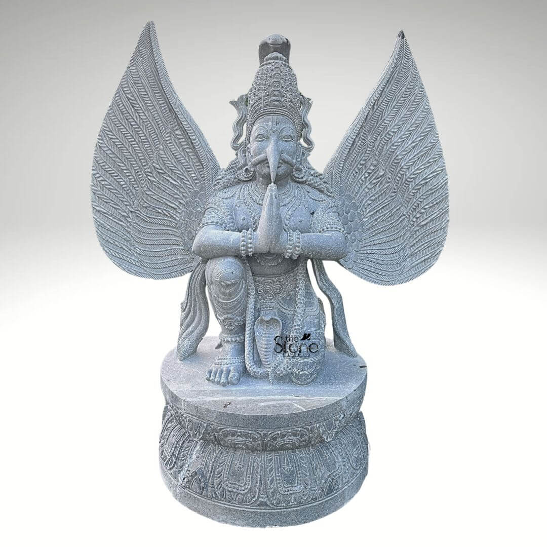 Grey Stone Garuda Statue 5.5ft 6 A detailed front-facing photograph captures a magnificent large Garuda stone idol, standing 5.5 feet tall. The statue is meticulously hand-carved from a single block of natural grey stone presenting the mythical bird-deity Garuda in a reverent kneeling posture. Garuda is positioned on a robust circular lotus-carved pedestal base, his hands pressed together in the Anjali Mudra a gesture of prayer and devotion held near his chest. His face is carved with sharp defined features including an eagle-like beak piercing eyes and a traditional crown with intricate patterns all rendered in deep relief that highlights the skill of the artisan. A defining feature of this large grey stone Garuda idol is its massive fully spread wings which rise dramatically behind him mirroring his height. Each wing feather is individually and delicately carved creating a texture that is both visually detailed and structurally impressive conveying a sense of power and flight. Garuda's body is adorned with finely carved traditional ornaments including layered necklaces armbands bracelets and a sacred thread all contributing to his divine appearance. A coiled serpent is visibly carved beneath one of his feet further enhancing the traditional iconography associated with this celestial mount of Lord Vishnu. This five-and-a-half-foot large Garuda stone idol is an exceptional example of traditional Indian stone carving, making it a perfect centerpiece for any article discussing grand stone sculptures or Hindu mythology. Its commanding size and detailed craftsmanship make it suitable for prominent placement in a home temple a garden sanctuary as an architectural element in a larger space or within a cultural institution. The choice of durable grey stone ensures its longevity making it a timeless investment for spiritual and aesthetic appreciation. The image clearly displays the unique iconographic elements and the high level of artistry that define such a premium stone idol.