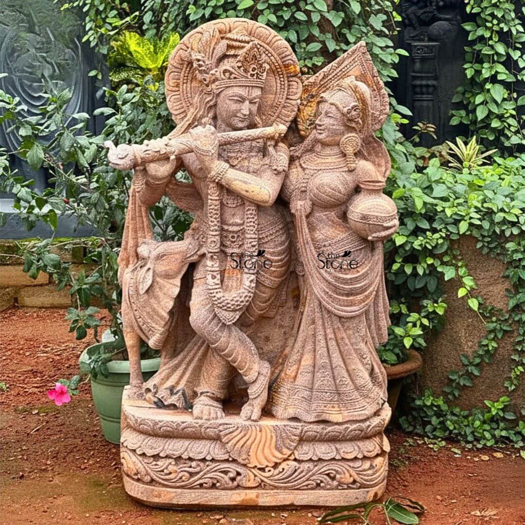 Radha Krishna Stone Statue with Cow 4ft 3 A magnificent 4-foot Large Radha Krishna Statue in Brown Sandstone stands gracefully in a lush outdoor garden setting radiating divine love and harmony. The statue carved from warm reddish-brown sandstone depicts Lord Krishna and Radha in a loving standing pose. Krishna distinguished by his elaborate crown and serene expression holds a flute to his lips poised as if playing a melodious tune with his head gently tilted towards Radha. Radha stands gracefully beside him adorned with a similar crown and traditional attire her gaze fixed on Krishna while she carries a pot of water or butter symbolizing her devotion and nurturing spirit. This detailed carving beautifully captures their divine romance and pastoral setting. The craftsmanship is exquisite with intricate details defining their elaborate crowns delicate jewelry and flowing traditional garments. The sandstone's natural texture and subtle variations in color enhance the antique feel of the sculpture bringing out the tender expressions on their faces and the intricate patterns of their attire. A halo-like circular aureole with ornate patterns frames Krishna's head further emphasizing his divine presence. A small docile cow is subtly depicted near Krishna's feet symbolizing his role as a protector and caretaker of cattle adding to the pastoral charm. The large scale of the 4-foot statue allows for an impressive level of detail and presence. The entire ensemble rests on a multi-tiered intricately carved pedestal adorned with lotus petal designs and other traditional motifs providing a stable and dignified foundation. The statue is surrounded by vibrant green plants and foliage creating a natural and harmonious backdrop. A single pink flower is visible near the base of the statue adding a delicate touch of color. This Large Radha Krishna Statue in Brown Sandstone is an exquisite example of religious artistry designed to bring blessings love and aesthetic beauty to any home or garden making it an ideal focal point for devotion perfectly aligning with an article focusing on such charming and grand Radha Krishna statues.