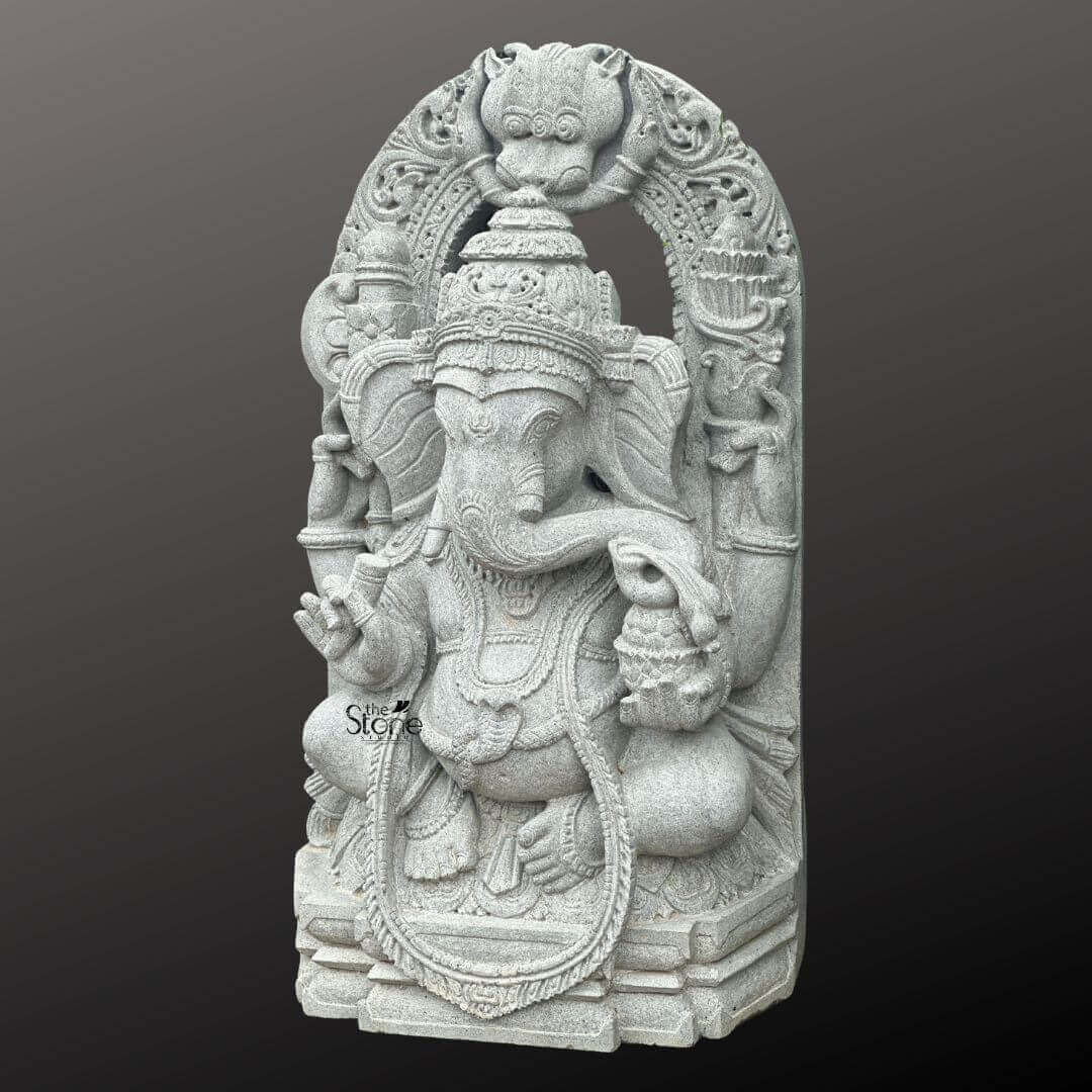 Ganesha Statue with Makara Torana 3ft 5 A magnificent 3-foot Lord Ganesha Stone Statue intricately carved from what appears to be a light grey stone stands powerfully against a dark, gradient background. The statue depicts Lord Ganesha in a regal seated posture exuding benevolence wisdom and an aura of divine grace. His iconic elephant head is finely sculpted complete with large ears expressive eyes and his distinctive broken tusk delicately poised to write or interact. He is adorned with an elaborate crown multiple layers of traditional jewelry including necklaces armlets and a sacred thread draped across his chest all rendered with exceptional detail in the light grey stone. Ganesha is portrayed with four arms holding his customary attributes a modak in one hand an axe or goad in another and his lower right hand extended in a gesture of blessing offering reassurance and protection. The intricate details on his garments the patterns on his halo and the careful sculpting of his prominent pot belly are all clearly visible showcasing the high artistry of the carving. The light grey stone emphasizes these textures and details lending a serene and timeless elegance to the deity and suggesting a weathered ancient feel that speaks to its profound presence. The statue is dramatically framed by a grand ornate arch that features exquisite mythical creature motifs and intricate traditional carvings along its entire span and supporting pillars. The top of the arch features a Kirtimukha a fierce lion-like face a common protective motif. This entire elaborate structure rests on a multi-tiered base also carved from the same light grey stone, providing stability and enhancing its grand, temple-like presence. This 3-foot Lord Ganesha Stone Statue with its impressive arch is an exquisite example of religious artistry designed to bring blessings peace and aesthetic grandeur to any space perfectly aligning with an article focusing on such a revered Lord Ganesha sculpture.