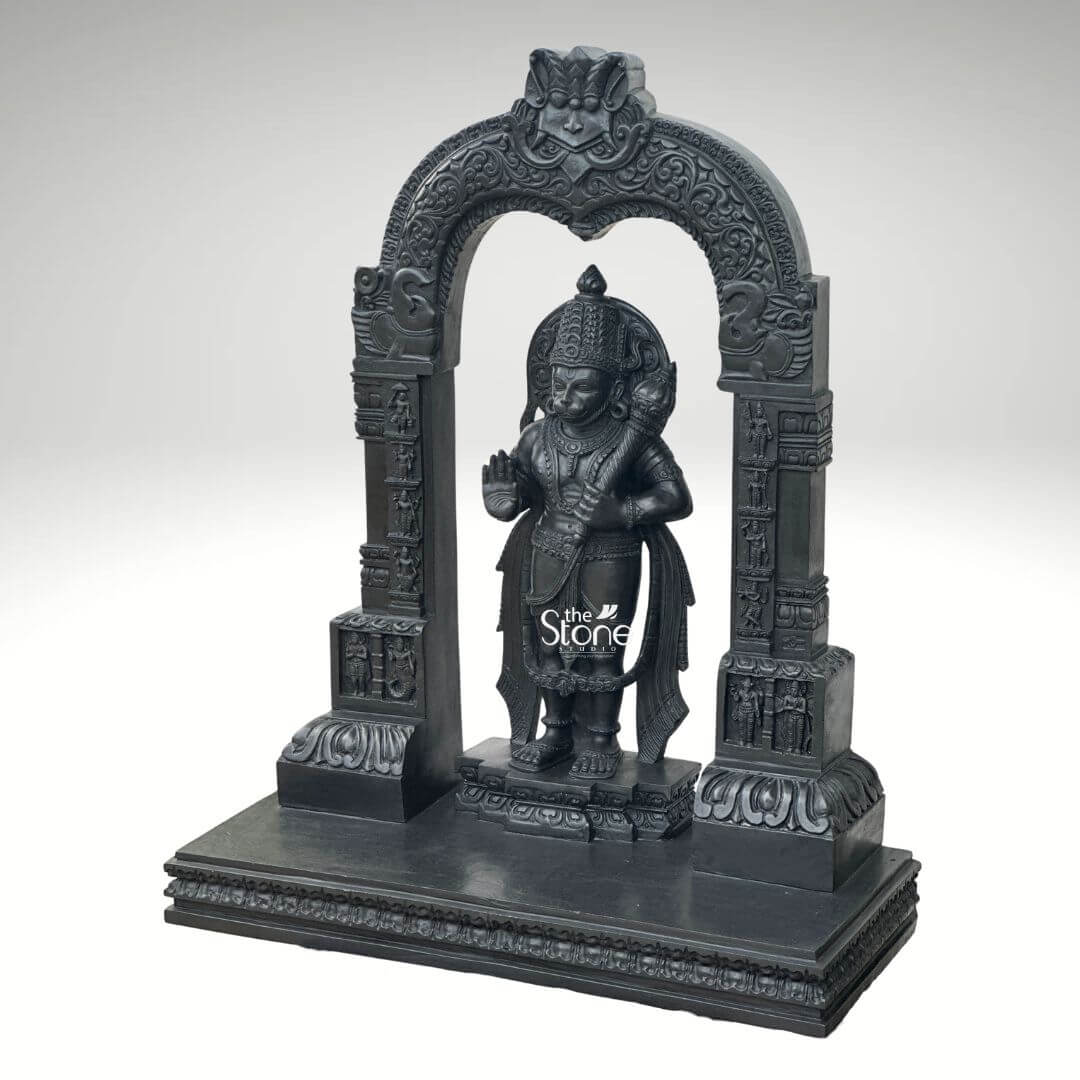 Black Stone Hanuman Statue with Arch 3ft 5 A commanding 3-foot Lord Hanuman Stone Statue with Gada intricately carved from deep black stone stands prominently within an ornate archway against a clean light grey background creating a powerful and reverent display. Lord Hanuman is depicted in a traditional standing posture radiating immense strength unwavering devotion and a vigilant readiness. He is adorned with an elaborate crown traditional jewelry around his neck chest and arms and finely sculpted flowing garments all rendered with exceptional detail in the lustrous dark stone. A central focus explicitly tied to the article's title is his left hand firmly grasping his formidable gada a potent symbol of his immense power valor and ability to overcome obstacles. His right hand is raised in a gesture of Abhaya Mudra offering fearlessness and divine protection to all devotees. The craftsmanship of this Blackstone Hanuman statue is truly outstanding with every minute detail meticulously brought to life. His facial features including his serene yet determined expression are precisely rendered conveying his spiritual resolve. The subtle depiction of his musculature beneath his garments hints at his legendary strength and agility. Lord Hanuman stands on a multi-tiered black stone pedestal which is seamlessly integrated into the broader base of the entire archway structure providing both stability and enhancing its grand temple-like presence. The statue is dramatically framed by a beautifully carved decorative arch also crafted from black stone. This arch features intricate traditional patterns floral motifs and smaller divine figures or mythological scenes meticulously carved into its supporting pillars and along the curve of the arch. These surrounding architectural elements elevate the spiritual grandeur and cultural richness of the entire piece reminiscent of ancient temple entrances. This impressive 3-foot Lord Hanuman Stone Statue with Gada and its integrated decorative arch serves as an ideal representation for an article focusing on such a divine sculpture symbolizing unwavering loyalty courage profound spiritual strength and the iconic weapon that defines much of his power.