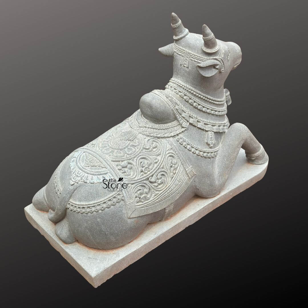 Nandikeshwara Stone Statue 45" 5 A high-angle rear three-quarter photograph captures a masterfully hand-carved Lord Shiva Nandi stone idol depicting the sacred bull in a traditional reclining posture. The statue is sculpted from a single dense block of dark grey blackstone which has been polished to a deep matte finish emphasizing the rich deep color and intricate sculptural details. Nandi is shown in a serene lying down position resting upon a simple elevated rectangular blackstone plinth. The view from the back reveals the powerful yet graceful form of the bull with careful attention paid to the musculature of the hindquarters and back. Nandi is richly adorned with traditional deeply carved ornaments that showcase the skill of the artisan. A large ornate blanket covers his back featuring elaborate symmetrical floral paisley and geometric patterns in high relief. This blanket extends down the sides with intricately carved fringed borders. Around his neck and hump he wears multiple layered necklaces of varying design including beaded chains bells and floral pendants all rendered with fine detail. The carving extends to his legs which are decorated with anklets. His horns are carefully shaped and point upwards and outwards. This Blackstone Nandi statue is an exceptional example of traditional South Indian temple carving art making it a perfect subject for an article discussing Lord Shiva Nandi stone idols. Its size detailed craftsmanship and traditional iconography make it suitable for placement in a home shrine a temple garden or as a central exhibit capturing the spiritual significance and cultural heritage of the deity. The durable blackstone material ensures the statue's longevity making it a timeless investment for both spiritual devotion and artistic appreciation. The photograph's clear focus on the statue's texture form and ornamentation provides a comprehensive view for the viewer.