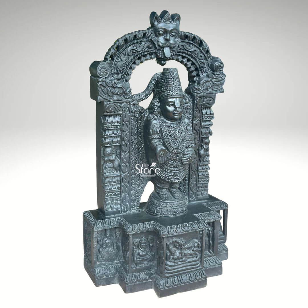 Sri Srinivasa Tirupati Balaji Statue 2.5ft 2 A detailed three-quarter view photograph presents a magnificent 2.5-foot-tall statue of Lord Srinivasa, also known as Tirupati Balaji meticulously hand-carved from a single block of dark dense blackstone. The statue depicts Lord Balaji standing in a samabhanga posture on a lotus pedestal which is itself supported by an ornate multi-tiered rectangular base. The dark stone has a deep matte finish that highlights the intricate sculptural details. Garuda the celestial eagle-mount is prominently carved at the apex of the elaborate prabhavali that frames the main deity. Below Garuda the arch is adorned with traditional floral and scrollwork motifs culminating in makara heads at the base of the pillars. Lord Balaji's divine form is richly adorned with finely carved traditional ornaments including multiple layered necklaces, elaborate armbands bracelets and anklets. His forehead bears the distinct Vaishnava U-shaped tilak mark and his eyes are half-closed in a serene benevolent expression. His upper right hand holds a discus while his upper left hand holds a conch shell. His lower right hand is raised in the Varada mudra and his lower left hand rests gracefully on his hip. The fine detail extends to his draped clothing which is textured to resemble silk fabric held by a jeweled girdle. The rectangular base beneath the main figure is a work of art in itself featuring intricate bas-relief panels on its visible front face. The central panel depicts the reclining form of Lord Ranganatha resting on the serpent Adisesha attended by consorts. This central scene is flanked by smaller, detailed carvings of sages devotees, and other deities within architectural niches. The entire sculpture, from the ornate pedestal to the top of the arch reflects the traditional South Indian Hoysala and Vijayanagara architectural styles. This 2.5-foot blackstone statue is an exceptional piece of devotional art perfect for a home shrine or as a central exhibit capturing the spiritual essence and cultural heritage of the Venkateswara Temple. The photograph's clear focus on the statue's form and texture provides a comprehensive view for the viewer.