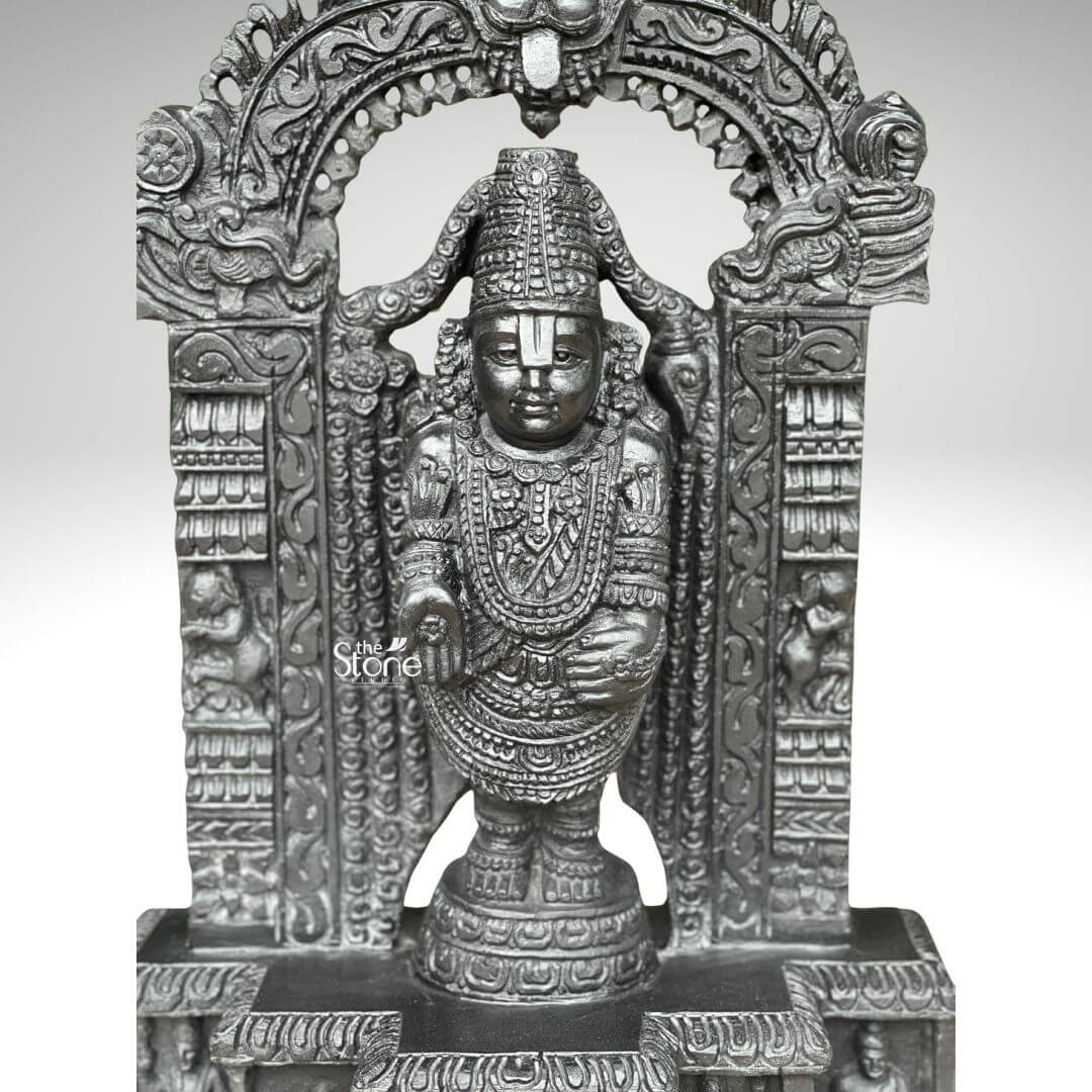 Sri Srinivasa Tirupati Balaji Statue 2.5ft 4 A front-facing photograph captures a meticulously hand-carved statue of Lord Venkateswara also known as Tirupati Balaji standing at 2.5 feet tall. The statue is sculpted from a single block of dark dense blackstone which has been polished to a deep matte finish that emphasizes the intricate sculptural details. The deity stands in a samabhanga posture upon a lotus pedestal which is itself supported by an ornate rectangular base. Garuda the celestial eagle-mount is prominently carved at the apex of the elaborate prabhavali that frames the main figure. Below Garuda the arch is adorned with traditional floral and scrollwork motifs culminating in makara heads at the base of the pillars. Lord Balaji's divine form is richly adorned with finely carved traditional ornaments including multiple layered necklaces, elaborate armbands bracelets, and anklets. His forehead bears the distinct Vaishnava U-shaped tilak mark and his eyes are half-closed in a serene benevolent expression. His upper right hand holds a discus while his upper left hand holds a conch shell. His lower right hand is raised in the Varada mudra and his lower left hand rests gracefully on his hip. The fine detail extends to his draped clothing which is textured to resemble rich, pleated silk fabric held by a jeweled girdle. The rectangular base beneath the main figure is a work of art in itself featuring intricate bas-relief panels on its visible front face. The central panel depicts the reclining form of Lord Ranganatha resting on the serpent Adisesha attended by consorts. This central scene is flanked by smaller detailed carvings of sages devotees and other deities within architectural niches. The entire sculpture from the ornate pedestal to the top of the arch reflects the traditional South Indian Hoysala and Vijayanagara architectural styles. This 2.5-foot blackstone Tirupati Balaji statue is an exceptional piece of devotional art perfect for a home shrine or as a central exhibit capturing the spiritual essence and cultural heritage of the Venkateswara Temple. The photograph's clear focus on the statue's form and texture provides a comprehensive view for the viewer.