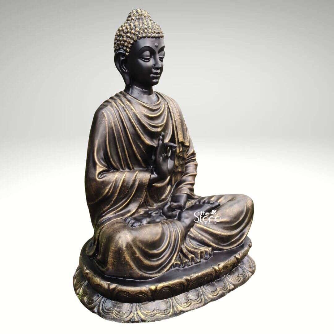Large Fibre Buddha Statue 5ft 4 A 5 feet meditating Buddha statue in fibre is shown from a three-quarter side angle against a plain light grey background allowing the sculpture’s posture finish and detailing to be clearly visible. The Buddha is seated cross-legged on a broad lotus pedestal with one hand raised near the chest in a calm teaching or blessing gesture and the other hand resting gently in the lap. The face has a peaceful meditative expression with softly closed eyes smooth facial features elongated earlobes and detailed curled hair highlighted in a golden tone. The statue has a rich black and antique gold finish that gives the fibre Buddha sculpture a classic elegant and spiritual appearance. Flowing robe folds are carefully sculpted across the shoulders arms chest and legs with gold accents enhancing the layered drapery and carved contours. The lotus base features repeated petal patterns around the lower edge adding symbolic meaning and decorative depth to the seated Buddha form. The angled view also shows the rounded shape of the pedestal the balanced body proportions and the graceful curve of the meditative pose. This image is well suited for an article about a Meditating Buddha Statue in Fibre because it clearly highlights the large 5 feet size lightweight fibre material antique finish, and serene spiritual presence of the sculpture. The statue can be imagined as a striking focal point for meditation rooms living areas entrance spaces yoga studios wellness centres garden corners or peaceful home décor settings. With its calm expression lotus seat raised hand gesture and refined black-gold finish this 5 feet Buddha fibre statue represents mindfulness peace elegance and timeless devotional beauty.