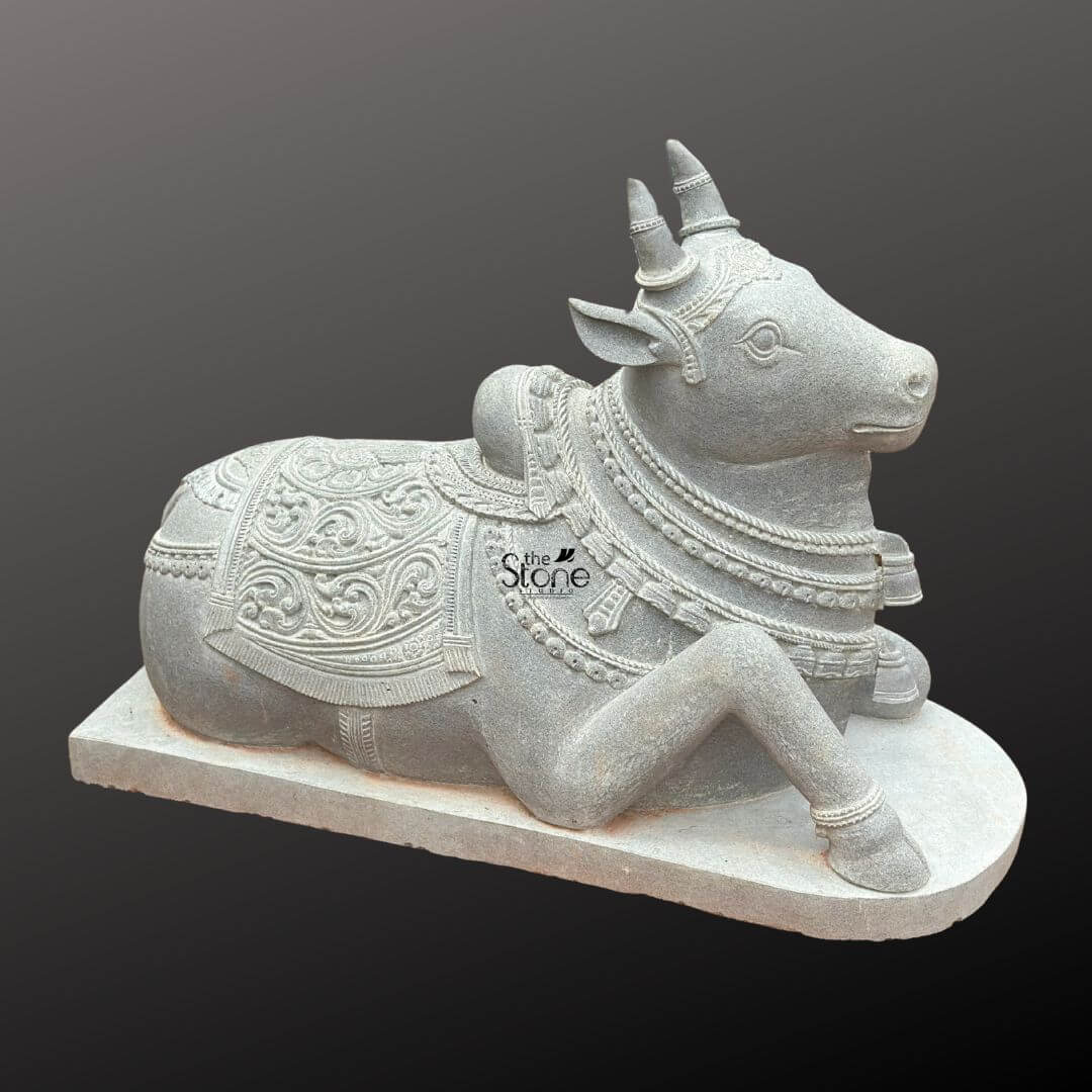 Nandikeshwara Stone Statue 45" 1 A three-quarter view photograph presents a masterfully hand-carved Nandikeshwara stone statue depicting the divine bull Nandi the sacred mount of Lord Shiva. The statue is sculpted from a single solid block of dark grey blackstone which has been polished to a deep matte finish emphasizing the intricate sculptural details. Nandi is shown in a traditional, serene reclining posture resting on a simple elevated rectangular blackstone plinth. His head is alert and turned slightly to his right gazing with a calm benevolent expression. The fine-grained texture of the stone is visible giving the piece an authentic ancient feel. Nandi is richly adorned with traditional deeply carved ornaments that showcase the skill of the artisan. He wears multiple layered necklaces of varying designs including beaded chains and bell-shaped pendants along with a decorative forehead band and ear ornaments. A large ornate blanket with elaborate floral paisley and geometric patterns in high relief covers his back and hindquarters extending down the sides with fringed borders. The carving extends to his legs which are decorated with anklets and his horns and humps are carefully rendered. The overall form is powerful yet graceful capturing the spiritual and aesthetic essence of Nandi. This Blackstone Nandi statue is an exceptional example of traditional South Indian temple carving art making it a perfect subject for an article discussing Nandikeshwara stone statues. Its size detailed craftsmanship and traditional iconography make it suitable for placement in a home shrine a temple garden or as a central exhibit capturing the spiritual significance and cultural heritage of the deity. The durable blackstone material ensures the statue's longevity making it a timeless investment for both spiritual devotion and artistic appreciation. The photograph's clear focus on the statue's texture form and ornamentation provides a comprehensive view for the viewer.