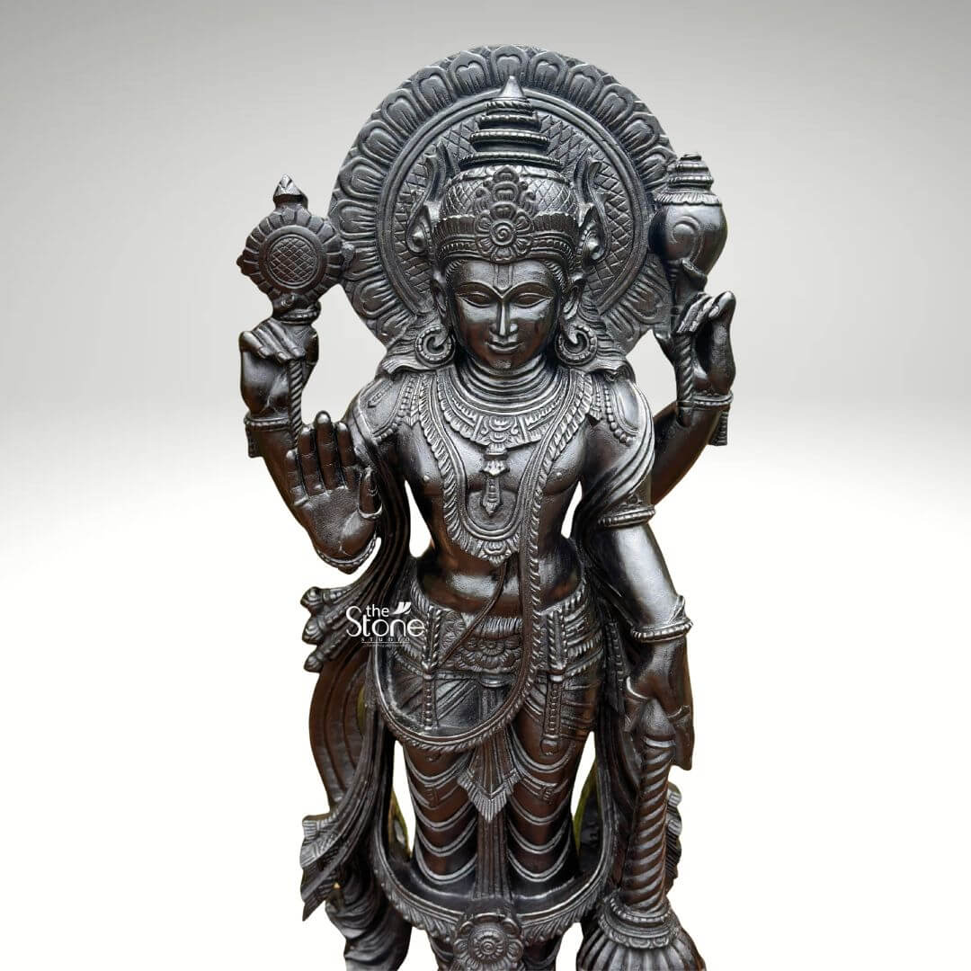 Black Stone Narayana Swamy Statue 3.5ft 7 A detailed close-up photograph captures the serene upper body and face of a majestic forty-one-inch statue of Lord Narayana Swamy meticulously hand-carved from a single block of deep black granite. The deity's face conveys a profound sense of inner peace and divine compassion with half-closed meditative eyes and a gentle benevolent smile. Its head is adorned with an intricately carved towering mukuta featuring detailed patterns a central floral motif and a small aureole all defined with exceptional precision against the dark stone. The fine natural crystalline structure and rich dark matte-to-satin finish characteristic of high-quality black granite are evident throughout the sculpture. The four-armed form of Lord Narayana is depicted with elaborate shallow-relief carvings of traditional South Indian temple jewelry including multiple necklaces armlets bracelets and a sacred thread all rendered with exceptional detail. The deity's four arms radiate outwards from the central figure showcasing the artisan's skill in defining each attribute. The upper right hand holds the chakra while the upper left hand holds the shankha. The lower right hand is shown in the abhaya mudra a gesture of fearlessness and blessing and a small portion of the gada is visible in the lower left hand. The statue conveys a sense of monumental presence and spiritual majesty with deep relief carvings defining the folds of the garments and the texture of the ornaments against the smooth skin. A grand intricately carved prabhavali forms a halo behind the deity adorned with stylized floral and geometric patterns. The statue is presented against a clean neutral graduated grey background ensuring the focus remains entirely on the detailed figure of Narayana Swamy and its artistic craftsmanship. The black granite material ensures durability and a timeless quality making this large statue a commanding presence for any devotional space.