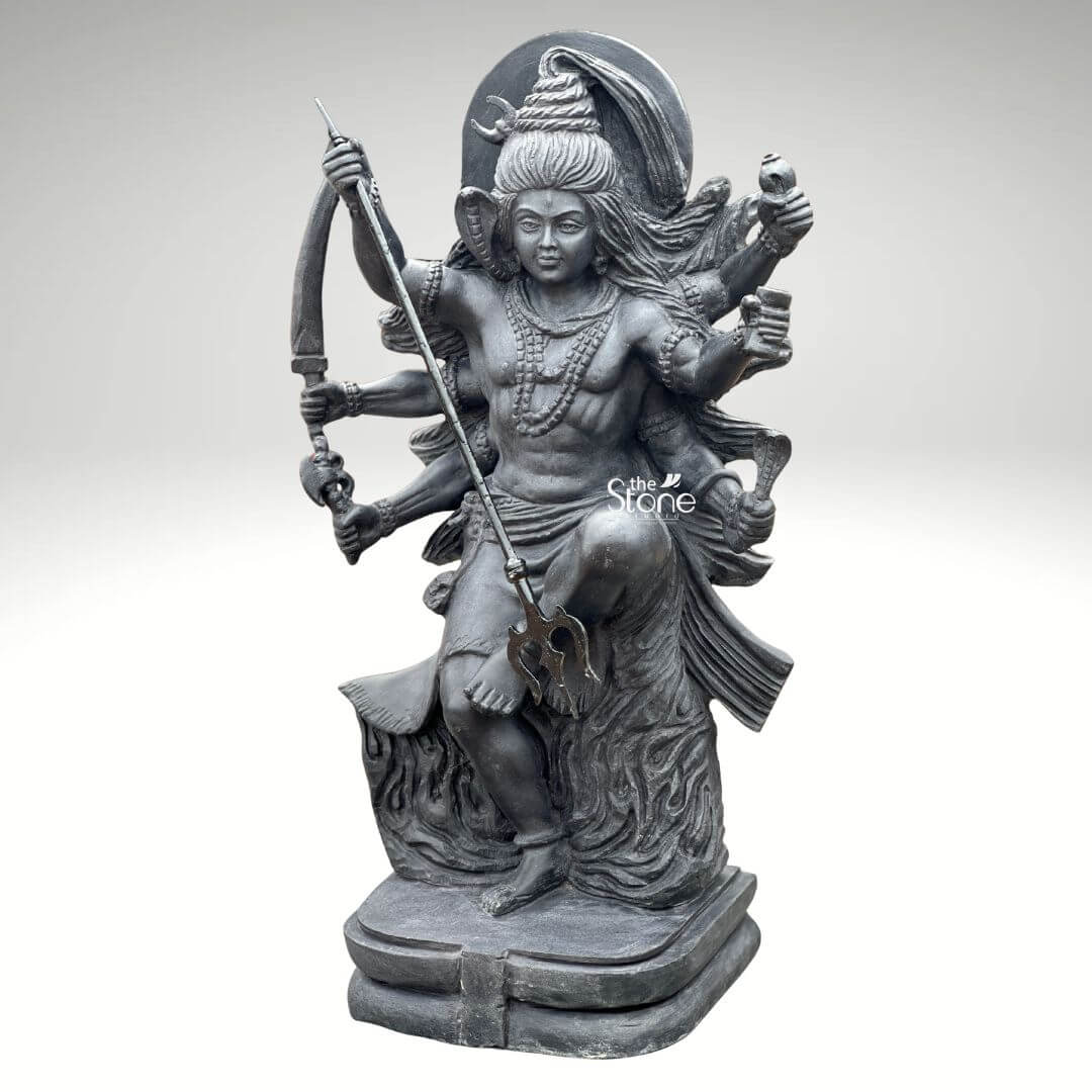 2 Feet Tandava Shiva Statue 4 A full-body studio photograph of a magnificent 2-foot tall Nataraja Shiva Statue masterfully hand-carved from a single block of dense polished black stone. The statue captures Lord Shiva in the dynamic and powerful Ananda Tandava pose the dance of cosmic bliss, set upon a multi-tiered lotus pedestal. Shiva is depicted with ten arms fanned out to convey movement holding various divine attributes including a trident and damaru drum. His matted hair is piled high adorned with a crescent moon and a serpent and flares out dramatically emphasizing the energy and rhythm of his celestial dance. The intricate details of the carving from the delicate folds of Shiva's garment to the fierce yet serene expression on his face and the form of the demon Muyalakan subdued beneath his foot showcase the exquisite craftsmanship involved in creating this handcrafted piece. The black stone used for the statue has a deep rich hue with a subtle waxy luster giving it a powerful and reverent presence that complements the traditional iconography. The ten arms are arranged with precision some raised in gestures of protection and blessing others holding attributes like a conch shell a fire pot and a scripture. Shiva's right leg is raised bent at the knee while his left leg is planted firmly on the demon Muyalakan who lies subdued beneath his foot symbolizing the triumph of knowledge over ignorance. The dynamism of the dance is conveyed through the arching of his body and the intricate arrangement of his limbs creating a sense of controlled power and divine ecstasy. This 2-foot black stone Nataraja Shiva statue stands as a perfect embodiment of Indian temple art and a meaningful centerpiece for any sacred space or home altar. Its compact size allows for placement where its intricate details and spiritual energy can be admired. The dark monolithic stone gives it a timeless quality, while the powerful iconography of the Tandava pose evokes a sense of cosmic rhythm creation and destruction making it an ideal choice for anyone seeking to bring a touch of Eastern spirituality and artistic heritage into their environment. The photograph presents the statue against a clean neutral white background ensuring the focus remains entirely on the form texture and spiritual significance of the black stone sculpture.