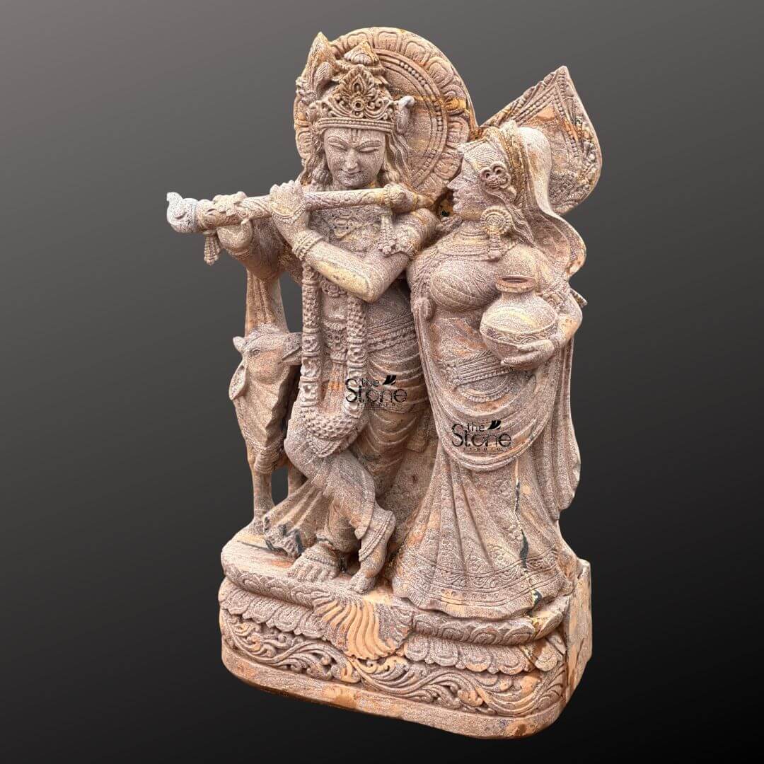 Radha Krishna Stone Statue with Cow 4ft 7 A magnificent 4-foot Natural Brown Sandstone Radha Krishna Murti intricately carved stands gracefully against a dark gradient background radiating divine love and harmony. The statue carved from warm reddish-brown sandstone prominently showcases its natural brown hue and texture depicting Lord Krishna and Radha in a loving standing pose. Krishna distinguished by his elaborate crown and serene expression holds a flute to his lips poised as if playing a melodious tune. Radha stands gracefully beside him adorned with a similar crown and traditional attire her gaze fixed on Krishna while she carries a pot of water or butter symbolizing her devotion and nurturing spirit. This detailed carving beautifully captures their divine romance and pastoral setting. The craftsmanship is exquisite with intricate details defining their elaborate crowns delicate jewelry and flowing traditional garments. The sandstone's prominent natural texture and subtle variations in color enhance the antique feel of the sculpture bringing out the tender expressions on their faces and the intricate patterns of their attire. A halo-like circular aureole with ornate patterns frames Krishna's head further emphasizing his divine presence. A small docile cow is subtly depicted near Krishna's feet symbolizing his role as a protector and caretaker of cattle adding to the pastoral charm. The entire ensemble rests on a multi-tiered intricately carved pedestal adorned with lotus petal designs and other traditional motifs providing a stable and dignified foundation. The depth and detail of the carving across the entire 4-foot statue are remarkable showcasing the artistry of the piece and the beauty of the natural sandstone. This Natural Brown Sandstone Radha Krishna Murti is an exquisite example of religious artistry designed to bring blessings, love and aesthetic beauty to any home or temple making it an ideal focal point for devotion perfectly aligning with an article focusing on such charming and symbolically rich natural sandstone Radha Krishna statues.
