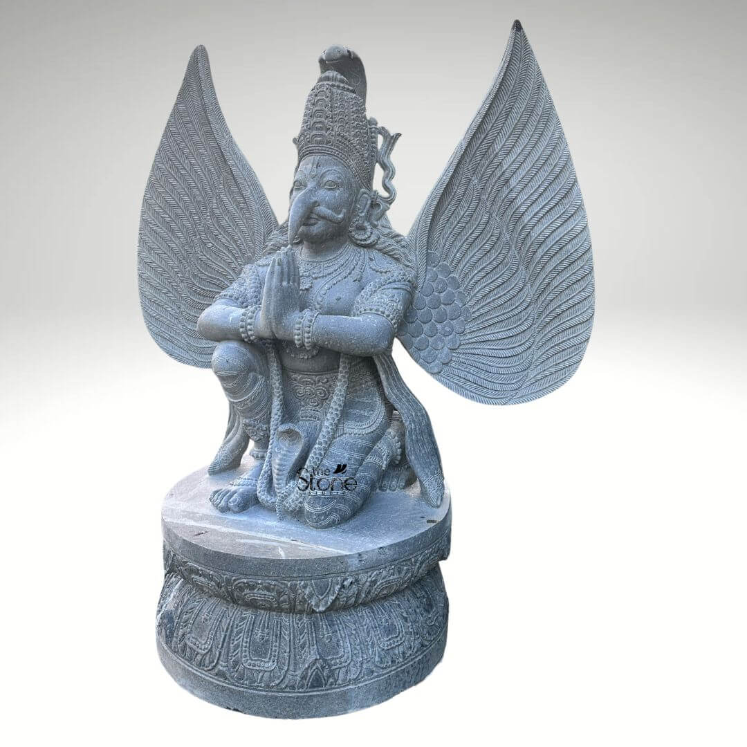 Grey Stone Garuda Statue 5.5ft 7 A detailed front-facing photograph captures a magnificent large Garuda stone idol standing 5.5 feet tall. The statue is meticulously hand-carved from a single block of natural grey stone presenting the mythical bird-deity Garuda in a reverent kneeling posture. Garuda is positioned on a robust circular lotus-carved pedestal base his hands pressed together in the Anjali Mudra a gesture of prayer and devotion held near his chest. His face is carved with sharp defined features including an eagle-like beak piercing eyes and a traditional crown with intricate patterns all rendered in deep relief that highlights the skill of the artisan. A defining feature of this large grey stone Garuda idol is its massive fully spread wings which rise dramatically behind him mirroring his height. Each wing feather is individually and delicately carved creating a texture that is both visually detailed and structurally impressive conveying a sense of power and flight. Garuda's body is adorned with finely carved traditional ornaments including layered necklaces armbands bracelets and a sacred thread all contributing to his divine appearance. A coiled serpent is visibly carved beneath one of his feet further enhancing the traditional iconography associated with this celestial mount of Lord Vishnu. This five-and-a-half-foot large Garuda stone idol is an exceptional example of traditional Indian stone carving, making it a perfect centerpiece for any article discussing grand stone sculptures or Hindu mythology. Its commanding size and detailed craftsmanship make it suitable for prominent placement in a home temple a garden sanctuary as an architectural element in a larger space or within a cultural institution. The choice of durable grey stone ensures its longevity making it a timeless investment for spiritual and aesthetic appreciation. The image clearly displays the unique iconographic elements and the high level of artistry that define such a premium stone idol.