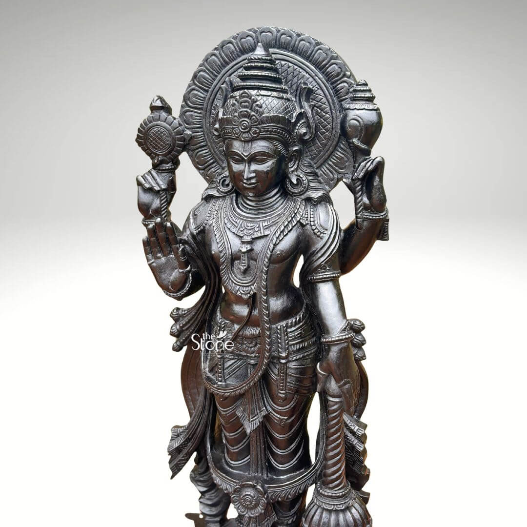 Black Stone Narayana Swamy Statue 3.5ft 8 A detailed full-body photograph captures a majestic forty-one-inch-tall statue of Lord Narayana Swamy meticulously hand-carved from a single solid block of deep black stone presenting a commanding devotional figure. The deity is depicted in a traditional standing posture upon an intricately carved circular lotus pedestal which features detailed petal layers and a decorative base. Narayana is shown with four arms radiating divine grace and power making it an ideal sacred art piece for a home temple or place of worship. The statue exhibits a rich dark matte-to-satin finish characteristic of high-quality natural black stone which absorbs light and highlights the depth and complexity of the carvings. The four-armed form of Lord Narayana holds traditional symbolic attributes each intricately defined against the dark textured stone. His upper right hand holds the chakra while his upper left hand holds the shankha. His lower right hand is shown in the abhaya mudra a gesture of fearlessness and blessing and his lower left hand rests gracefully holding the gada. The deity is adorned with elaborate shallow-relief carvings of traditional South Indian temple jewelry including multiple necklaces armlets bracelets, a sacred thread and a decorative mukuta all rendered with exceptional precision against the smooth stone. The statue conveys a sense of profound spiritual calm and majesty with the intricate details of his flowing dhoti with elaborate folds the detailed garlands and the serene facial expression all contributing to its devotional appeal. A grand intricately carved prabhavali forms a halo behind the deity adorned with stylized floral and geometric patterns that frame his head and shoulders. The statue is presented against a clean neutral graduated grey background ensuring the focus remains entirely on the detailed figure of Narayana Swamy and its artistic craftsmanship perfectly illustrating its suitability as a devotional focal point. The natural stone material ensures durability and a timeless quality.