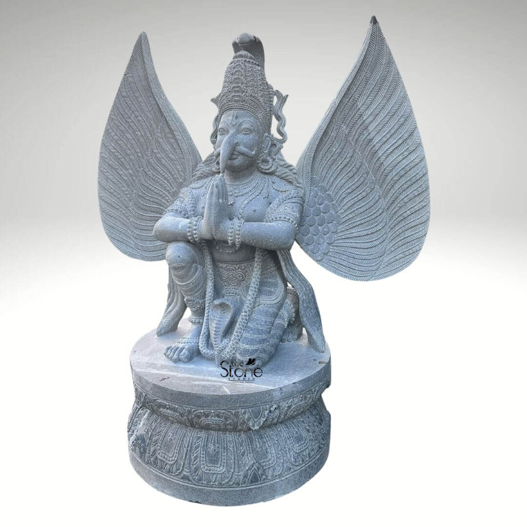 Grey Stone Garuda Statue 5.5ft 8 A front-facing full-length photograph captures a majestic 5.5-foot-tall Outdoor Garuda Statue meticulously hand-carved from a single block of natural grey stone. The statue depicts the mythical bird-deity Garuda in a reverent kneeling posture positioned upon a robust circular lotus-carved pedestal base. His hands are pressed together in the Anjali Mudra a gesture of prayer and devotion held near his chest. Garuda has a powerful human-like torso with an eagle-like face defined by a prominent beak piercing eyes and a traditional crown with intricate detailed patterns. His body is adorned with finely carved traditional ornament including layered necklaces armbands bracelets and a sacred thread all rendered in deep relief that highlights the skill of the artisan. A striking feature of this outdoor Garuda statue is its massive fully spread wings which rise dramatically behind him mirroring his height and creating a sense of divine presence. Each wing feather is individually and delicately carved creating a texture that is both visually detailed and structurally impressive. The grey stone itself possesses a natural slightly matte finish with subtle crystalline variations giving the sculpture an authentic ancient feel and underscoring the natural quality of the material, which is ideal for enduring the elements outdoors. A coiled serpent is visibly carved beneath one of his feet further enhancing the traditional iconography associated with this celestial mount of Lord Vishnu. This five-and-a-half-foot grey stone statue is an exceptional example of traditional Indian stonework making it a perfect subject for an article discussing outdoor Garuda statues. Its commanding size and detailed craftsmanship make it suitable for prominent placement in a garden sanctuary a courtyard as an architectural element in a larger landscaped space or within a cultural institution. The choice of durable grey stone ensures its longevity making it a timeless investment for spiritual and aesthetic appreciation in an outdoor environment. The image clearly displays the unique iconographic elements and the high level of artistry that define such a premium stone idol.
