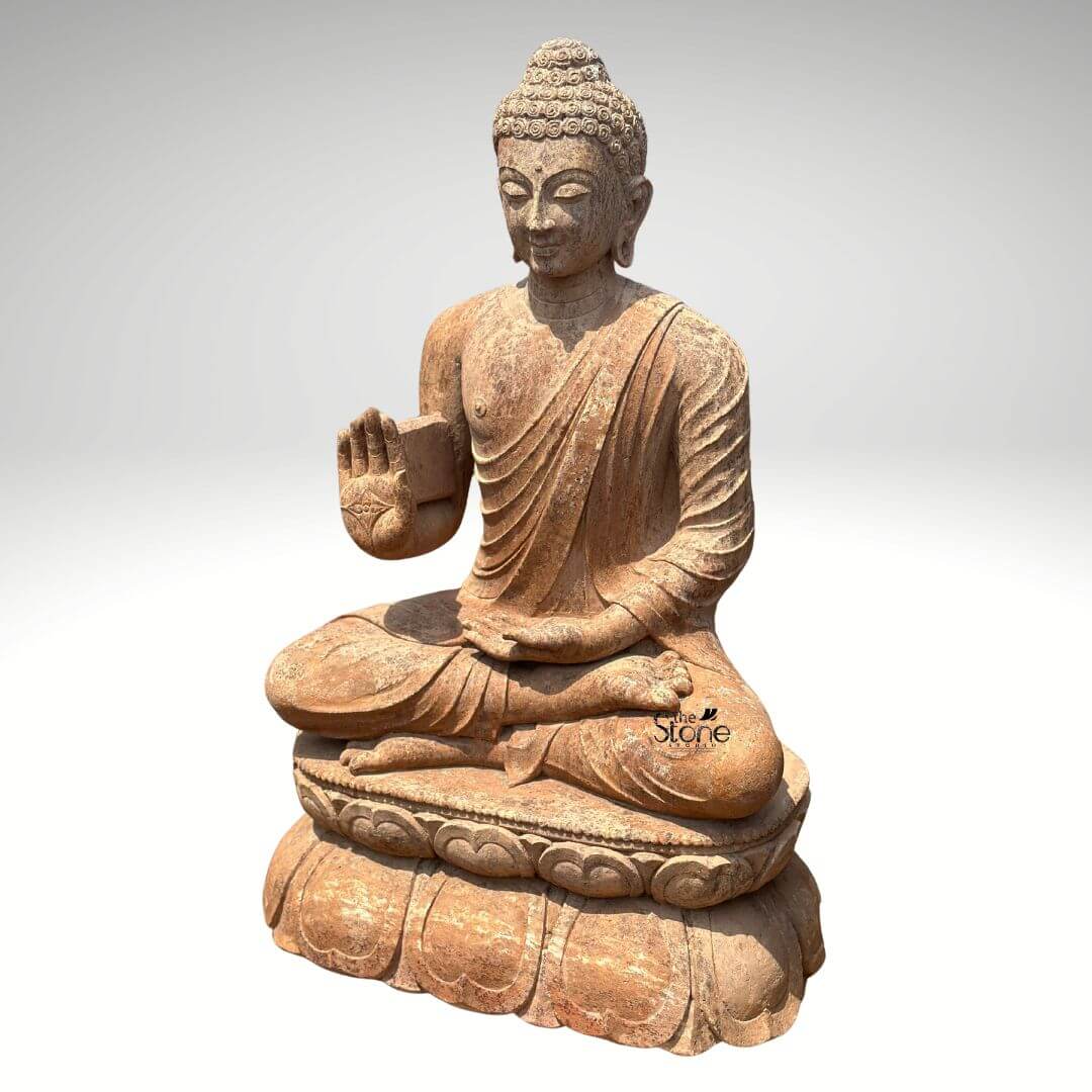 Sandstone Buddha Statue in Abhaya Mudra 3ft 2 A magnificent 3-foot Outdoor Sandstone Buddha Statue stands prominently against a clean light grey background radiating peace and reassurance. The statue carved from a warm reddish-brown sandstone depicts Buddha in a serene meditative posture seated in the full lotus position symbolizing deep concentration and spiritual enlightenment. His right hand is raised to shoulder height with the palm facing outwards fingers extended upwards forming the Abhaya Mudra the gesture of fearlessness blessing and protection. His left hand rests gently in his lap palm upwards signifying contemplation and receptivity. The natural earthy hue and weathered texture of the sandstone give the sculpture an authentic ancient feel making it perfectly suited for an Outdoor environment. The Buddha's facial features are exquisitely carved radiating profound peace and wisdom. His eyes are gently closed or cast downwards in contemplation and a subtle serene smile graces his lips. His head is adorned with tightly coiled hair culminating in the ushnisha the cranial bump symbolizing his supreme spiritual knowledge and enlightenment. The flowing drapery of his robes is sculpted with soft elegant folds emphasizing the texture of the sandstone and contributing to the statue's graceful and tranquil form. The rustic worn appearance of the sandstone enhances its charm and allows it to blend seamlessly into a natural garden or patio setting gaining character over time through exposure to the elements. The Buddha is seated upon a beautifully sculpted double-lotus pedestal with each petal intricately carved symbolizing purity spiritual growth and the emergence of enlightenment from the mundane. The robust and detailed base supports the statue with dignity making it a focal point. This 3-foot Sandstone Buddha statue with its clear depiction of the Abhaya Mudra and durable sandstone material is an exquisite example of religious artistry designed to bring blessings peace and aesthetic beauty to any outdoor space aligning perfectly with an article focusing on Outdoor Sandstone Buddha Statues.