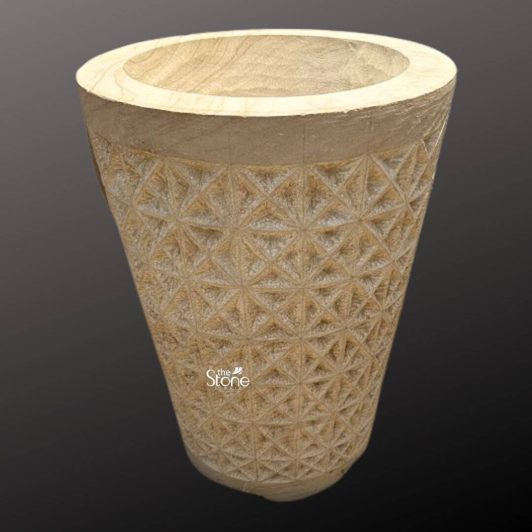 Natural Sandstone Planter Pot 2ft 4 A full-view photograph of a stunning 2-foot tall outdoor sandstone flower pot presented against a dark neutral gray gradient background that accentuates its form. The planter is hand-carved from a solid block of warm beige sandstone exhibiting a rich natural texture and tonal variations that highlight its stone origin. It is fashioned into a classic slightly tapered cylindrical shape with a thick smooth uncarved rim that provides a clean contrast to the body. The overall impression is one of rustic elegance and robust craftsmanship perfectly suited for enhancing any garden landscape. The central body of the sandstone planter pot is adorned with an intricate deeply incised geometric pattern. This pattern consists of a repeating tessellation of diamond and triangular facets creating a textured three-dimensional surface that catches the light and adds significant visual depth. The precision of the carving demonstrates a high level of artisanal skill turning the functional object into a piece of sculptural art. The natural porosity of the sandstone is clearly visible emphasizing its durability and suitability for outdoor environments where it can withstand the elements while allowing soil and plants to breathe. This substantial 2-foot stone planter pot is a versatile and attractive choice for any outdoor setting. Its scale and intricate design make it an ideal focal point for a patio deck or garden path providing a sturdy and stylish home for large flowering plants or small shrubs. The image clearly conveys the texture scale and elaborate geometric carving of the planter making it a perfect visual reference for an article discussing natural stone elements in garden design and the beauty of hand-carved sandstone products.