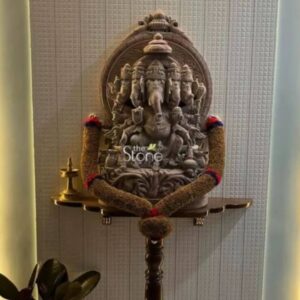 A three-quarter view photograph capturing a beautifully adorned 30-inch tall Panchmukhi Ganesha statue masterfully hand-carved from a light-colored soapstone. The statue depicts the five-headed form of Lord Ganesha with four heads visible facing different cardinal directions and one head looking upward all resting upon an elaborate multi-tiered ornate arch that serves as an aureole. The statue is seated in a graceful posture on a carved lotus pedestal with various attributes like an axe a modak and a lotus held in his multiple hands. The soapstone exhibits a natural warm tone and the intricate carvings showcase a high level of detail particularly in the crowns jewelry and the fine textures of the deity's garments. The Panchmukhi Ganesha statue is positioned centrally on a traditional dark wood wall-mounted shelf which has decorative carvings on its front edge. The statue itself is elegantly draped with a long colorful beaded garland made of brown red and blue segments that encircle the figure. To the left of Ganesha on the shelf rests a small polished brass oil lamp with a traditional design adding a warm ritualistic touch to the shrine. The shelf is mounted against a textured off-white wall with vertical paneling creating a sophisticated background. Below the shelf the top of a dark wooden stand is visible holding the entire structure. This soapstone Panchmukhi Ganesha statue stands as a captivating centerpiece for a home temple radiating a sense of divinity and peace. The soft ambient light from the nearby walls accentuates the rich textures of the soapstone carving and the warm glow of the brass diya. The placement of the statue on the traditional wooden shelf combined with the ritual elements evokes a deep sense of reverence and spiritual connection. This significant piece of Indian religious art is a perfect representation of the devotion and craftsmanship found in Mumbai making it an ideal choice for anyone seeking a sacred focal point in their home. The statue's scale and intricate details make it a truly remarkable object of contemplation.