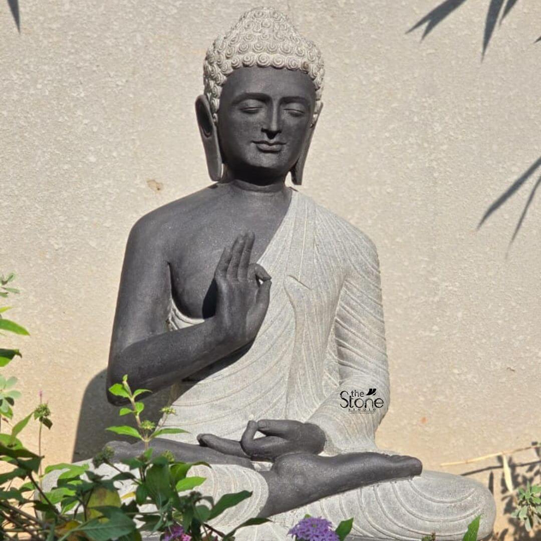 Two Tone Buddha Stone Statue for Home 3ft 3 A serene hand-carved Blackstone Buddha statue sits in a traditional meditative pose within a tranquil garden nook embodying the essence of peace. The three-foot-tall statue showcases a striking two-tone finish with the Buddha's body and head carved from a deep dark blackstone exhibiting a smooth polished texture that highlights the rich dark tones. This contrasts beautifully with his monastic robes which are rendered in a contrasting lighter grey or off-white stone featuring a subtle granular texture. This distinct separation emphasizes the form of the robes as they drape over his crossed legs and shoulders adding depth and visual interest to the sacred art. The Buddha is depicted seated with his legs crossed in a full lotus position on a simple integrated dark blackstone base nestled amidst lush greenery. His hands are gracefully positioned in the vitarka mudra symbolizing discussion and the transmission of teachings with the right hand raised and the thumb and index finger forming a circle while the left hand rests in his lap. His facial expression is calm and contemplative with downcast eyes a gentle smile and tightly curled hair leading to a cranial ushnisha all rendered with precise craftsmanship that honors traditional iconography. The overall composition evokes a sense of deep tranquility and spiritual harmony. This three-foot Blackstone Buddha statue is an ideal illustration for an article discussing a Peace Buddha Statue perfectly demonstrating its suitability as a spiritual and aesthetic centerpiece for any outdoor area. Its substantial size ensures it functions as a commanding focal point while the natural stone texture harmonizes beautifully with surrounding plant life. The traditional design brings a sense of sacred history and calm to any garden landscape while the durable blackstone construction ensures it is both a beautiful and a meaningful addition to the home garden inviting reflection and enhancing the peaceful atmosphere of its surroundings. The detailed carving and harmonious integration of elements highlight its value as a piece of sacred art in a modern landscape.