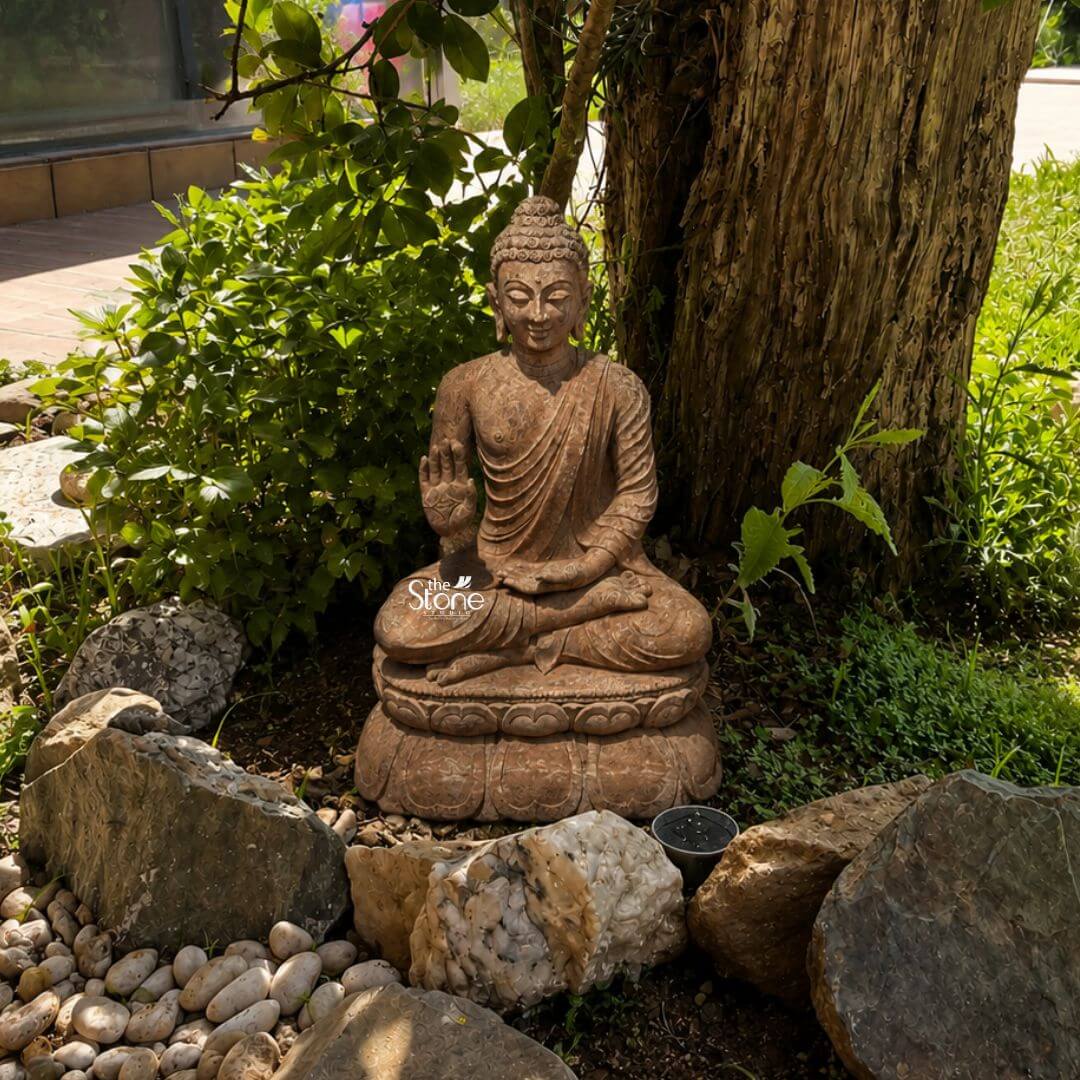 Sandstone Buddha Statue in Abhaya Mudra 3ft 3 A magnificent 3-foot Peaceful Sandstone Buddha Sculpture is beautifully situated in a natural garden setting nestled under the shade of a large tree creating an atmosphere of deep tranquility. The statue carved from a warm reddish-brown sandstone depicts Buddha in a serene meditative posture seated in the full lotus position symbolizing deep concentration and spiritual enlightenment. His right hand is raised to shoulder height with the palm facing outwards fingers extended upwards forming the Abhaya Mudra the gesture of fearlessness blessing and protection. His left hand rests gently in his lap palm upwards signifying contemplation and receptivity. The natural earthy hue and weathered texture of the sandstone lend the sculpture an authentic ancient feel perfectly blending with its organic surroundings. The Buddha's facial features are exquisitely carved radiating profound peace and wisdom with eyes gently closed or cast downwards in contemplation. A subtle serene smile graces his lips. His head is adorned with tightly coiled hair culminating in the ushnisha the cranial bump symbolizing his supreme spiritual knowledge and enlightenment. The flowing drapery of his robes is sculpted with soft elegant folds emphasizing the texture of the sandstone and contributing to the statue's graceful and tranquil form. The statue is placed on a beautifully sculpted double-lotus pedestal with each petal intricately carved symbolizing purity spiritual growth and the emergence of enlightenment from the mundane. The statue is thoughtfully surrounded by a ring of various natural rocks and smooth white pebbles creating a dedicated meditative space at the base of the tree. Lush green foliage including small bushes and ground cover fills the background and foreground further enhancing the natural harmony. The thick textured trunk of the tree provides a grand organic backdrop. This 3-foot Peaceful Sandstone Buddha Sculpture is an exquisite example of a garden deity designed to bring blessings peace and aesthetic beauty to any outdoor space aligning perfectly with an article focusing on such serene and contemplative stone sculptures.