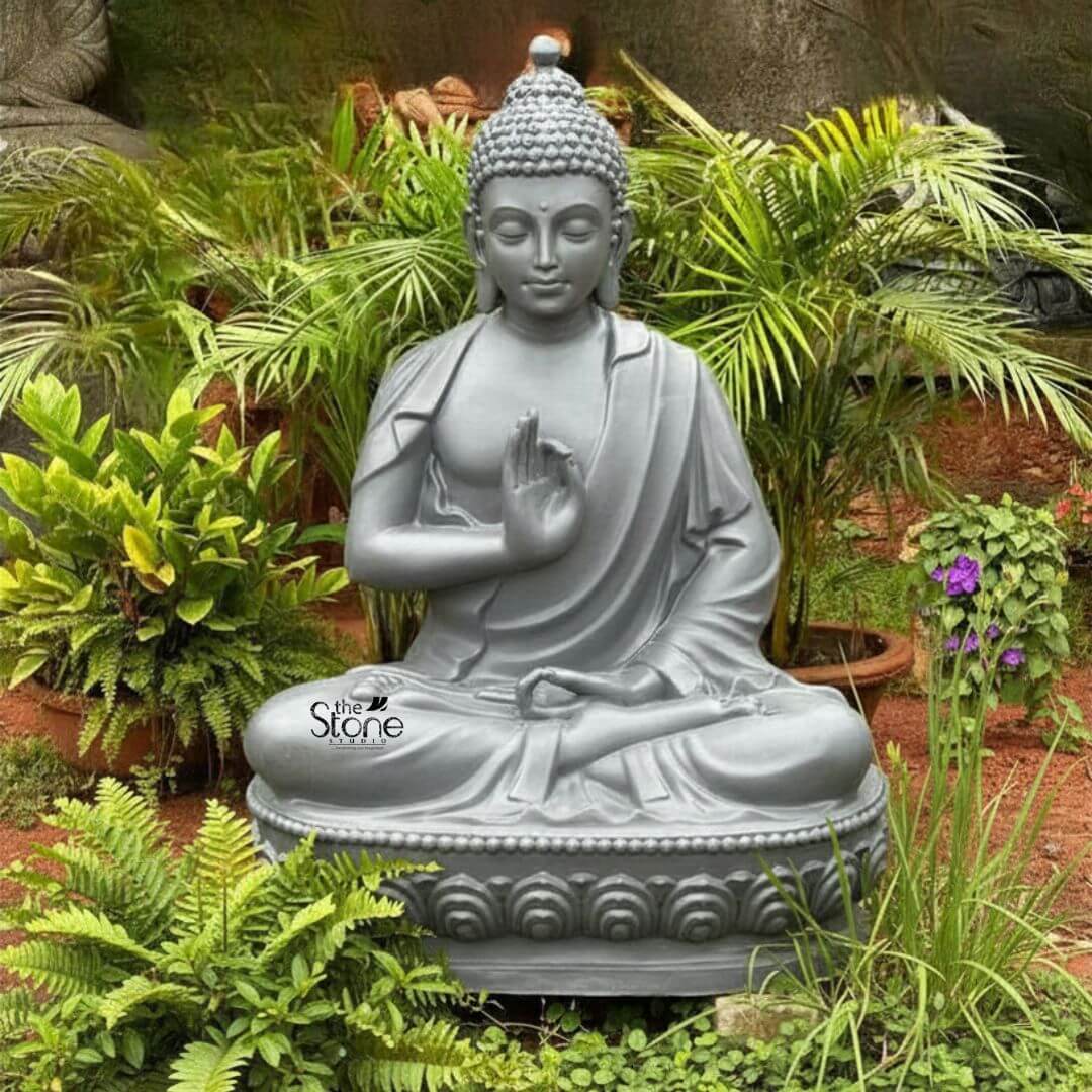 Seated Buddha Statue in Vitarka Mudra 3.5ft 3 A captivating 3.5-foot Peaceful Sitting Buddha Statue crafted from fiber and finished in a serene light grey is beautifully nestled amidst lush green foliage in what appears to be a garden or outdoor setting. The Buddha is depicted in the classic meditative full lotus position a posture symbolizing deep concentration and spiritual enlightenment. His right hand is raised in the Vitarka Mudra forming a circle with the thumb and index finger while the other fingers are extended signifying the transmission of teaching and intellectual discourse which contributes to a peaceful mind. His left hand rests gently in his lap palm upwards representing meditation and receptivity. The Buddha's face is carved with remarkable detail radiating profound peace and inner wisdom. His eyes are gently closed or downcast conveying a state of deep introspection and calm that aligns perfectly with the statue's peaceful description. A subtle, serene smile graces his lips and his elongated earlobes a traditional symbol in Buddhist iconography subtly reference his detachment from worldly possessions. His head is adorned with tightly coiled hair culminating in the ushnisha the symbolic cranial protuberance that signifies his supreme spiritual knowledge and enlightenment. The flowing drapery of his robes is expertly rendered with soft folds and graceful lines conveying the lightness and texture of fabric further enhancing the statue's tranquil and elegant form. The statue is seated upon a beautifully sculpted double-lotus pedestal with each petal intricately carved symbolizing purity spiritual growth and the emergence of enlightenment from the mundane. The natural backdrop of vibrant green ferns potted plants and hints of flowering purple plants creates a harmonious and organic environment for the Buddha emphasizing its role as a source of calm and serenity. This impressive 3.5-foot Buddha Fibre statue embodying profound spiritual meaning and artistic excellence is an ideal representation for a Peaceful Sitting Buddha Statue perfect for a home garden meditation space or any area designed to foster tranquility and contemplation.