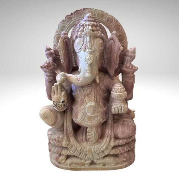 A front-facing studio photograph presents a twelve-inch tall Ganpati idol meticulously carved from a single piece of natural pink sandstone. The statue depicts the Hindu deity Ganesha seated in a relaxed posture with one leg folded and the other pendent resting upon a multi-tiered lotus-carved pedestal base. The stone exhibits a soft dusty pink hue with subtle natural variations in color and texture throughout its surface giving the piece an authentic artisanal quality. Ganesha has a serene expression with his trunk gently curving to his left holding a sweet modak. He is adorned with intricate deeply carved traditional jewelry including elaborate necklaces armbands anklets and a richly detailed crown that frames his head. The four-armed deity holds symbolic attributes in his hands, rendered with fine detail. His upper right hand holds a goad while his upper left hand holds a noose. His lower right hand is raised in the Abhaya mudra, a gesture of blessing and protection and his lower left hand holds a bowl overflowing with modaks. The carving extends to his garments which are textured to resemble pleated fabric held by an ornate girdle. A decorative prabhavali with floral and geometric patterns rises behind his head, completing the divine form. The entire sculpture is finished with a smooth polish that highlights the crystalline structure of the pink stone and the depth of the relief work. This twelve-inch pink stone Ganesha statue is an ideal choice for a home shrine or as a decorative accent. Its compact size makes it suitable for placement on a mantelpiece desk or in a small puja ghar bringing a sense of spiritual tranquility and aesthetic beauty to any living space. The warm pink color of the stone adds a gentle and inviting element to the decor while the detailed craftsmanship reflects a deep respect for traditional Indian iconography. The durable stone material ensures the idol's longevity making it a cherished piece for years to come.