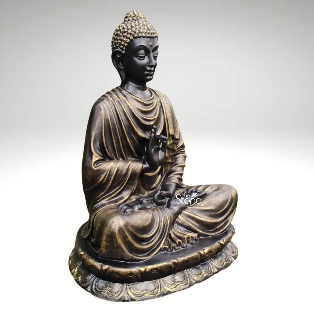 Large Fibre Buddha Statue 5ft 5 A premium 5 feet fibre Buddha sculpture is shown in a three-quarter side view against a clean light grey background highlighting its large size graceful seated form and detailed craftsmanship. The Buddha is seated cross-legged on a broad lotus pedestal with one hand raised near the chest in a symbolic teaching or blessing gesture and the other hand resting calmly in the lap. The face has a peaceful meditative expression with softly lowered eyes smooth facial features elongated earlobes and a composed posture that conveys serenity wisdom and spiritual balance. The statue features a striking black and antique gold finish giving the fibre Buddha sculpture a luxurious and timeless appearance. Golden highlights accent the curled hair robe folds and lotus base bringing depth and contrast to the dark surface. The robe is sculpted with flowing layered drapery across the shoulders chest arms and legs while the rounded lotus pedestal is decorated with repeated petal carvings that enhance the devotional and artistic appeal of the piece. The angled view clearly shows the dimensional form refined contours and premium finish of the large Buddha statue. This image connects well to an article about a Premium Fibre Buddha Sculpture by presenting the statue as a statement piece for spiritual and decorative spaces. The 5 feet Buddha fibre statue is suitable for meditation rooms entrance foyers living areas wellness centres yoga studios hotel interiors garden corners and peaceful décor settings where a large Buddha statue can create a calm focal point. Its serene expression antique gold detailing lotus base and elegant seated posture make it a beautiful choice for those seeking a premium Buddha sculpture that brings mindfulness sophistication and timeless spiritual charm into a space.