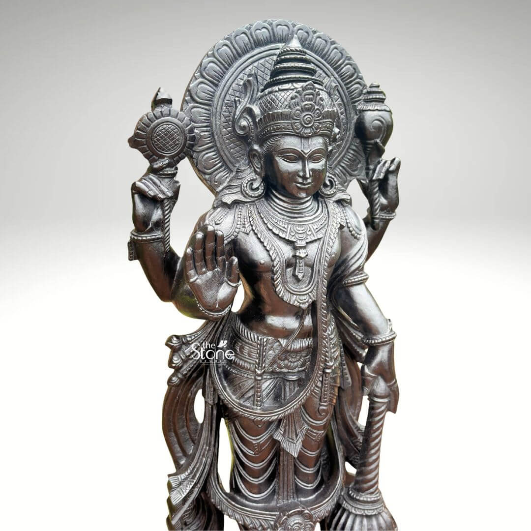 Black Stone Narayana Swamy Statue 3.5ft 9 A detailed full-body photograph captures a majestic forty-one-inch-tall statue of Lord Narayana Swamy meticulously hand-carved from a single solid block of deep black stone presenting a commanding devotional figure. The deity is depicted in a traditional standing posture upon an intricately carved circular lotus pedestal which features detailed petal layers and a decorative base. Narayana is shown with four arms radiating divine grace and power making it an ideal sacred art piece for a home temple or place of worship. The statue exhibits a rich dark matte-to-satin finish characteristic of high-quality natural black stone which absorbs light and highlights the depth and complexity of the carvings. The four-armed form of Lord Narayana holds traditional symbolic attributes each intricately defined against the dark textured stone. His upper right hand holds the chakra while his upper left hand holds the shankha. His lower right hand is shown in the abhaya mudra a gesture of fearlessness and blessing and his lower left hand rests gracefully holding the gada. The deity is adorned with elaborate shallow-relief carvings of traditional South Indian temple jewelry including multiple necklaces armlets bracelets a sacred thread and a decorative mukuta all rendered with exceptional precision against the smooth stone. The statue conveys a sense of profound spiritual calm and majesty with the intricate details of his flowing dhoti with elaborate folds the detailed garlands and the serene facial expression all contributing to its devotional appeal. A grand intricately carved prabhavali forms a halo behind the deity adorned with stylized floral and geometric patterns that frame his head and shoulders. The statue is presented against a clean neutral graduated grey background ensuring the focus remains entirely on the detailed figure of Narayana Swamy and its artistic craftsmanship perfectly illustrating its suitability as a devotional focal point. The natural stone material ensures durability and a timeless quality.